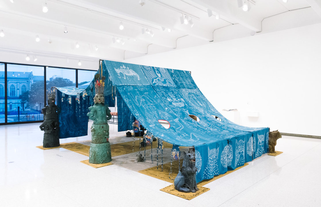 This photo of artist Candice Lin's installation features blue curtains that have been draped from poles, statues, and figures of cats.