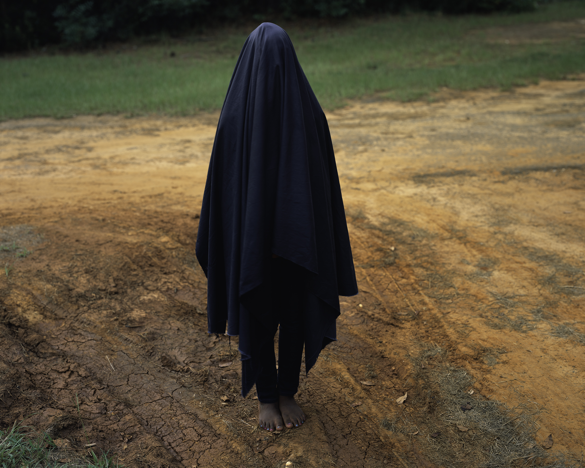 A person standing covered in all black barefoot on dirt with grass in the back