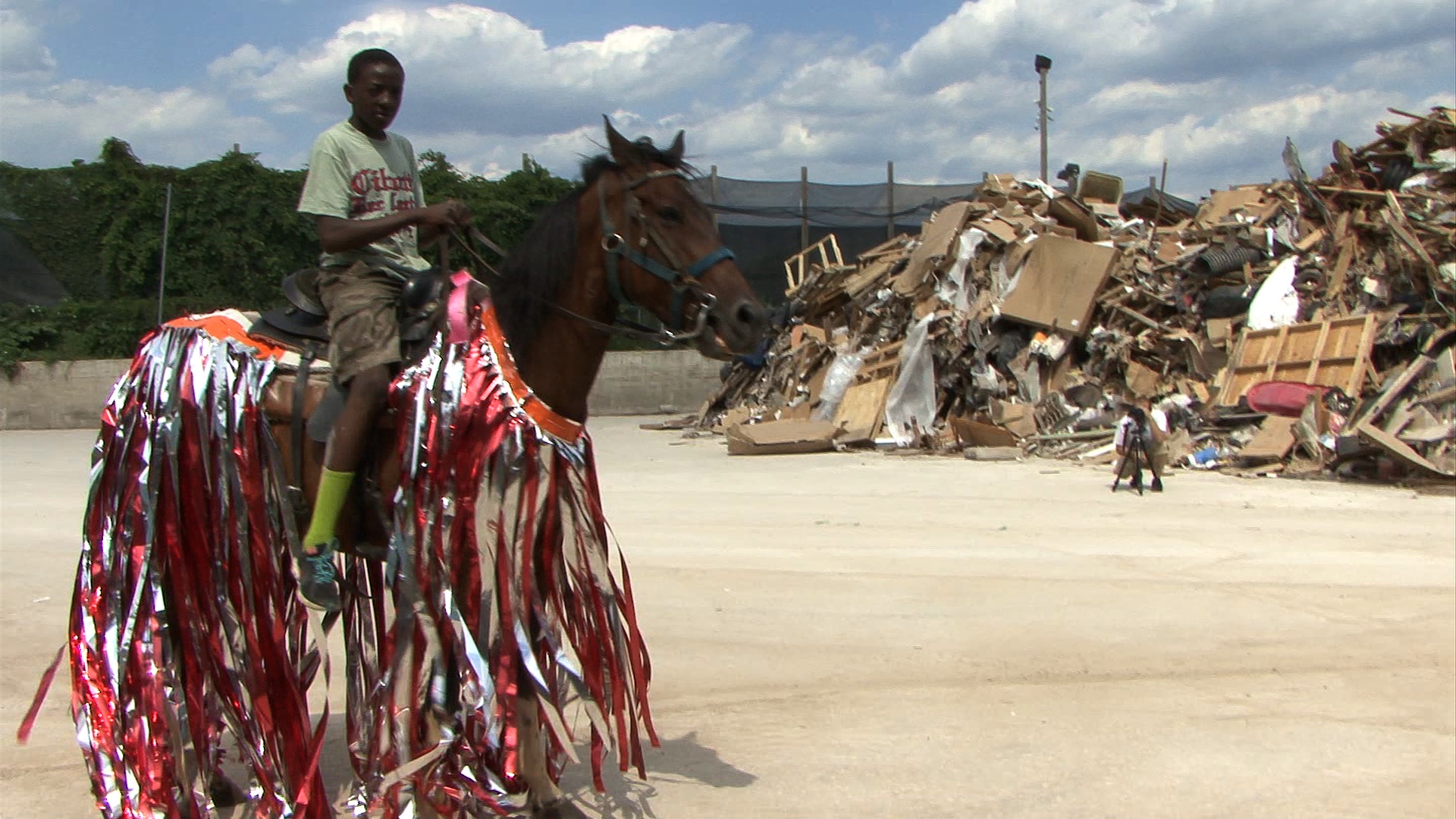 This is a photograph of a young boy riding a horse with read and silver ribbions hanging of of the horse in front of a mountain of discarded items taken by artist Mohamed Bourouissa