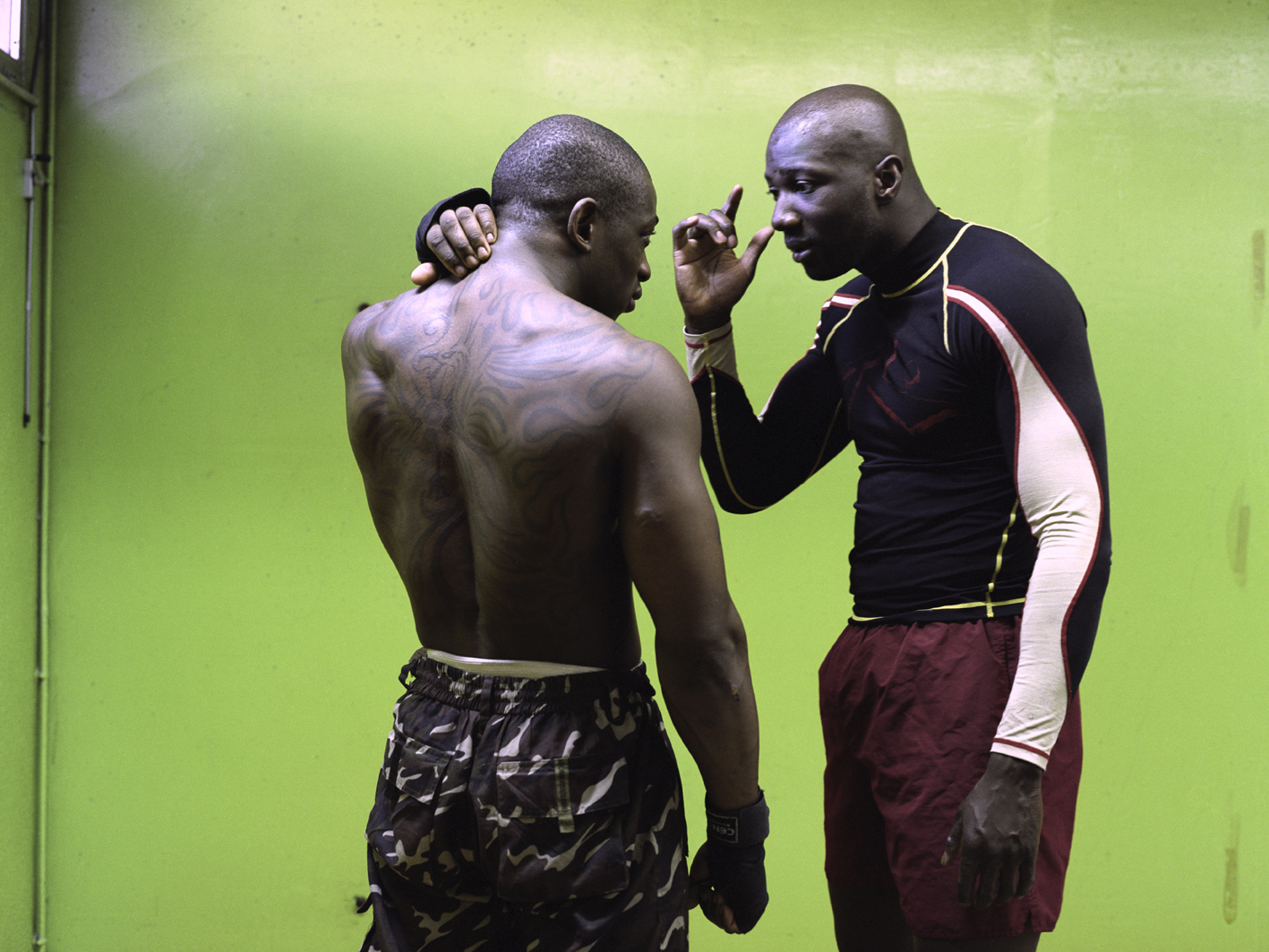 A photograph of two men talking in a room with green walls taken by artist Mohamed Bourouissa