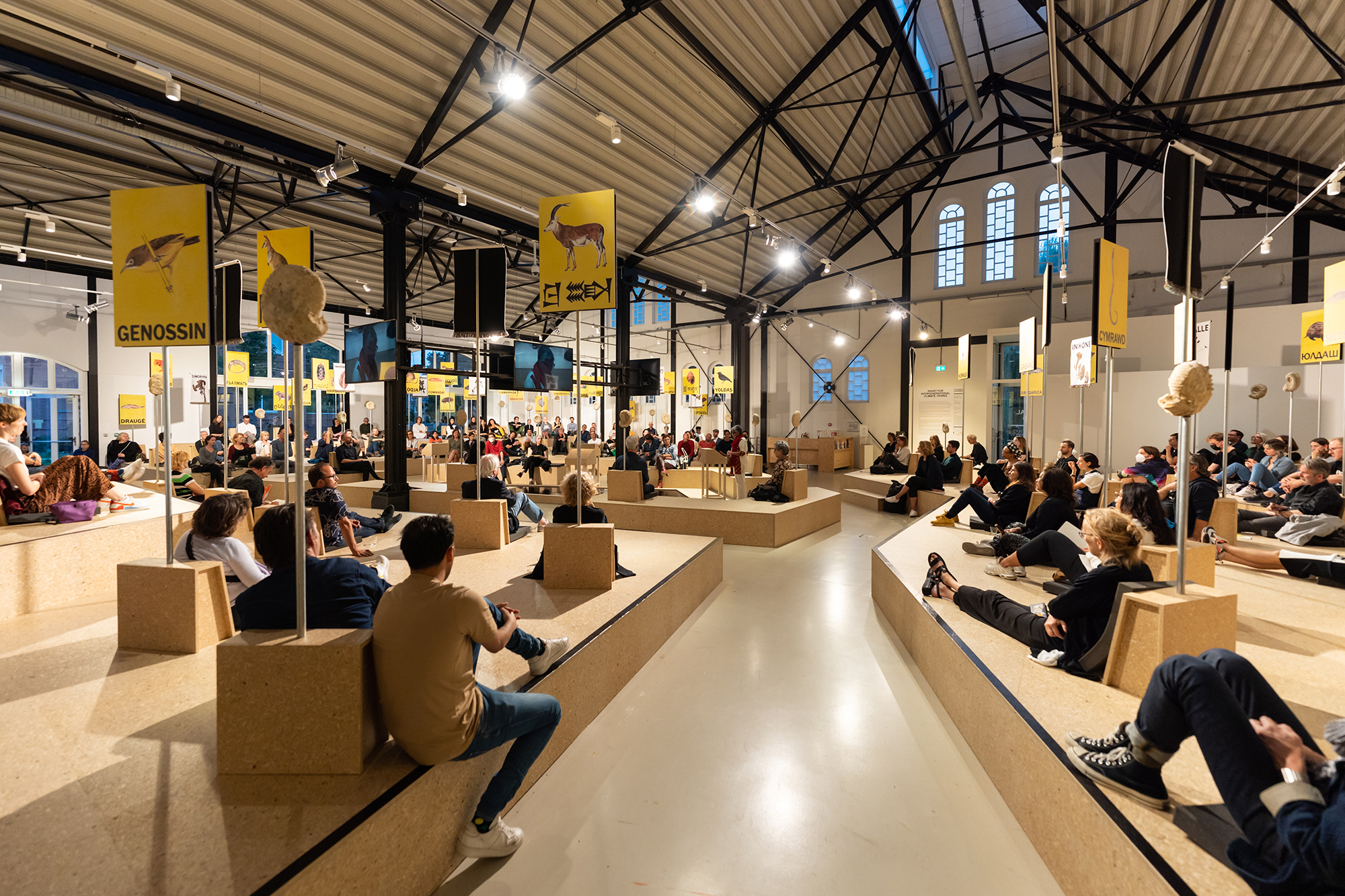 People gathered under in a large building filled with tan structures under yellow banners convening at the CICC.