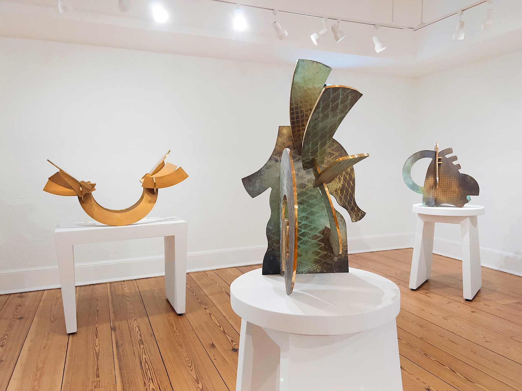 Green and gold bronze pieces rest atop white pedestals in the gallery space on top of an oaken wood floor.