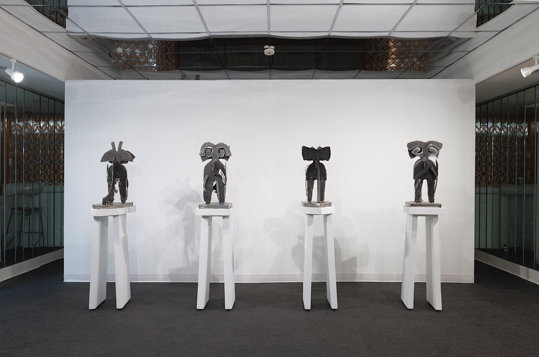 Four silver colored sculptures are lined up in a room of mirrors against a stark white background.