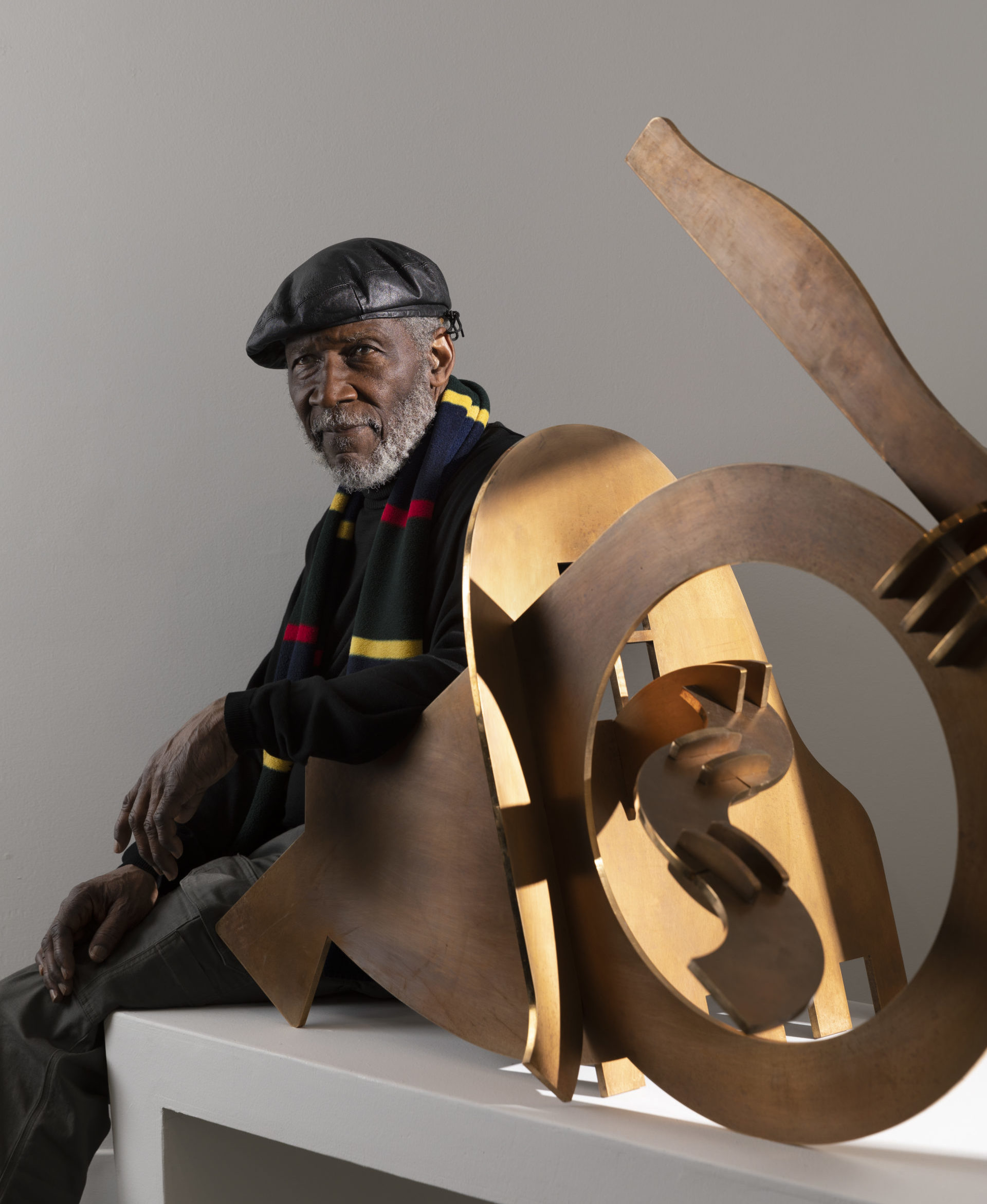 Curtis Patterson sits on a bench beside one of his bronze sculptures. He wears a black leather cap and a red and yellow striped scarf. He has a gray beard and a relaxed demeanor.