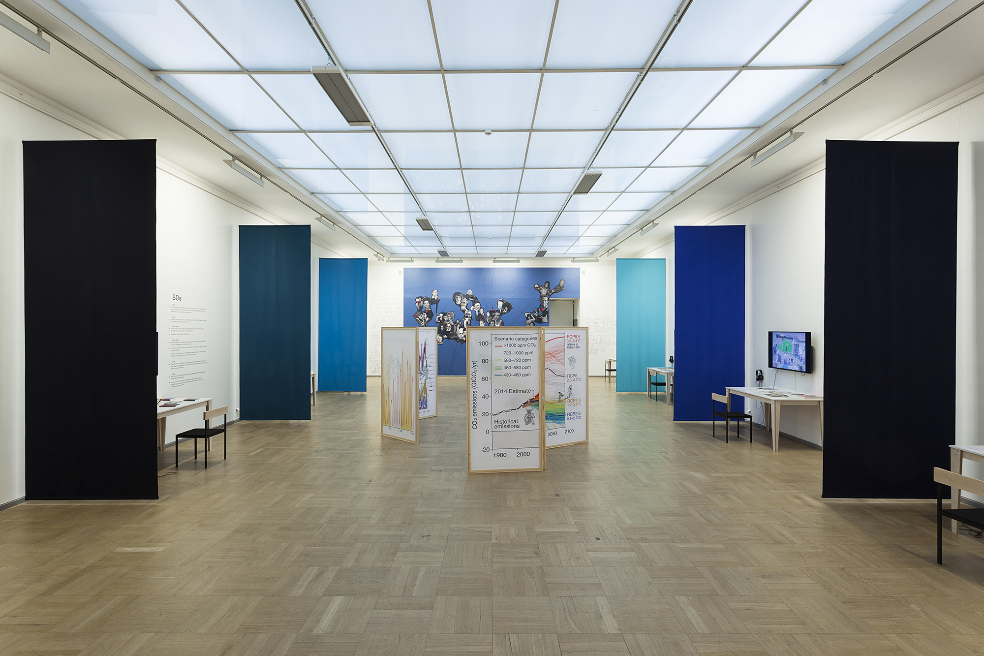 A bright room with blue dividers and a blue wall in the back with screens of graphs are arranged in the center.
