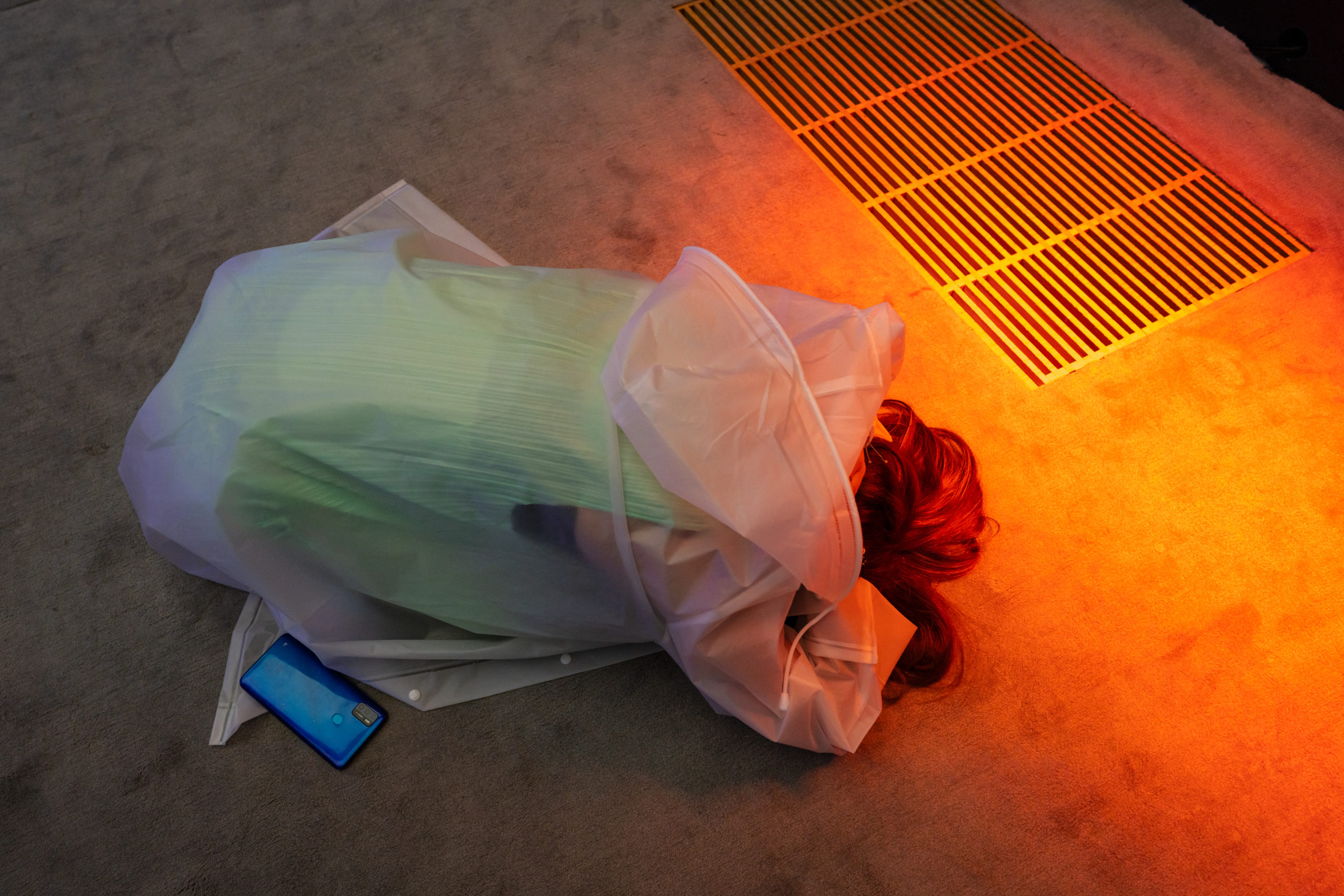 A person kneeling over with their head in their arms on the ground. A bright yellow orange light shines in the top right corner highlighting a vent on the floor next to the person and a bright blue phone on the ground in the bottom left corner. courtesy of the artist