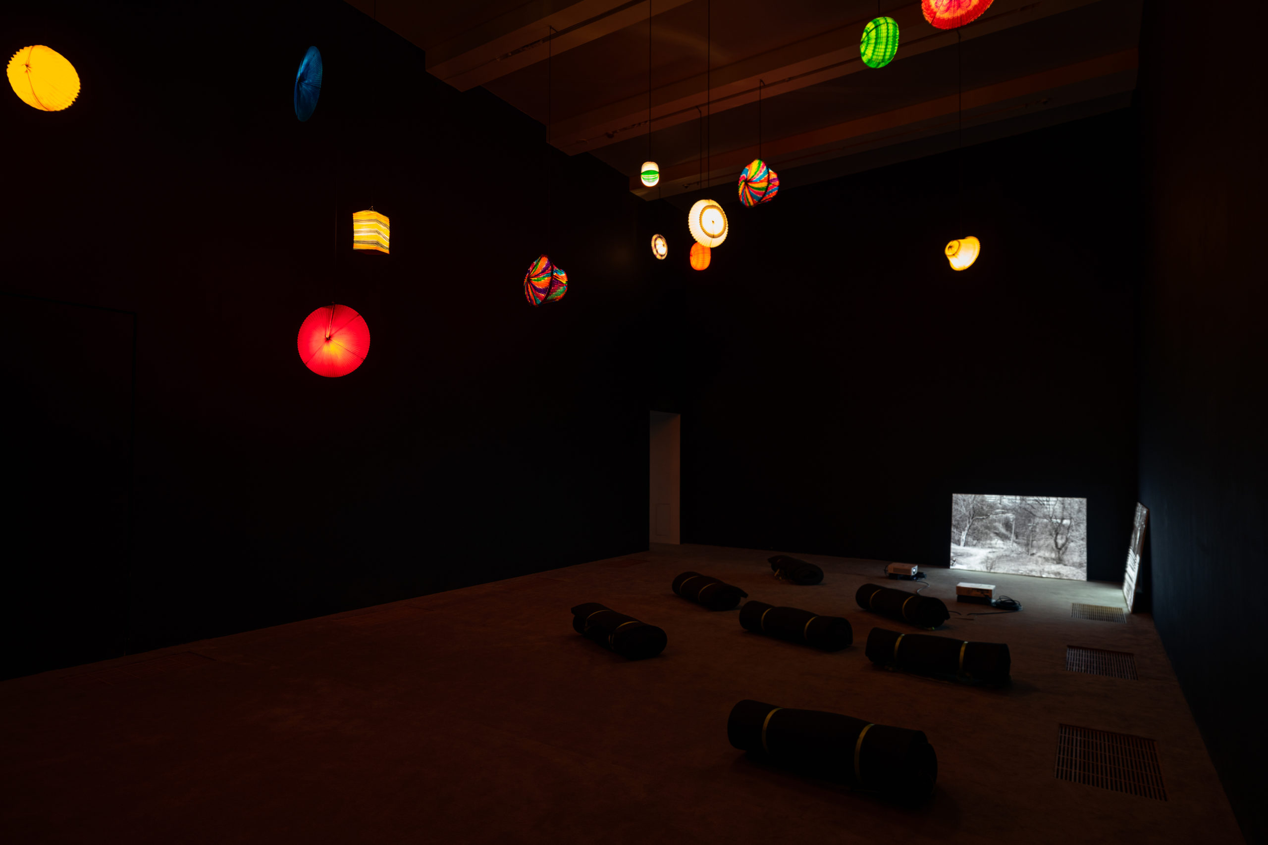 An image of a dark room with thirteen lanterns of different colors dimly lighting the room. Bottom right hand corner black and white images of the outside. Image provided by courtesy of artist.