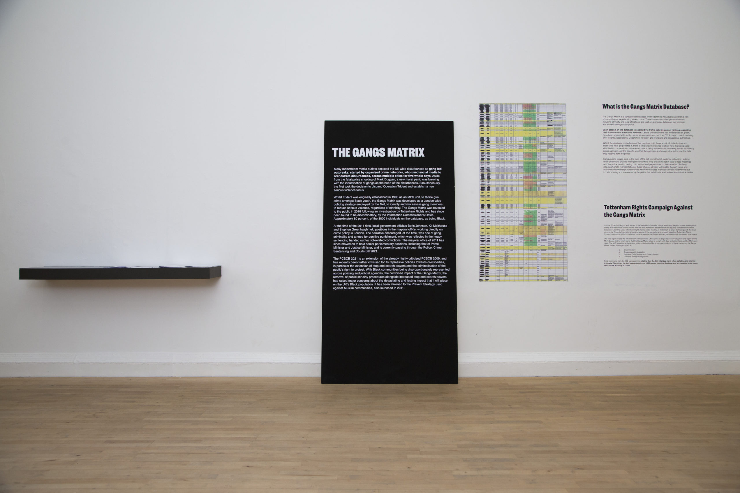 A black board with text about the Gangs Matrix stands against a white wall. To the left of it is a black bookshelf and to the right is a table and more text on the wall.