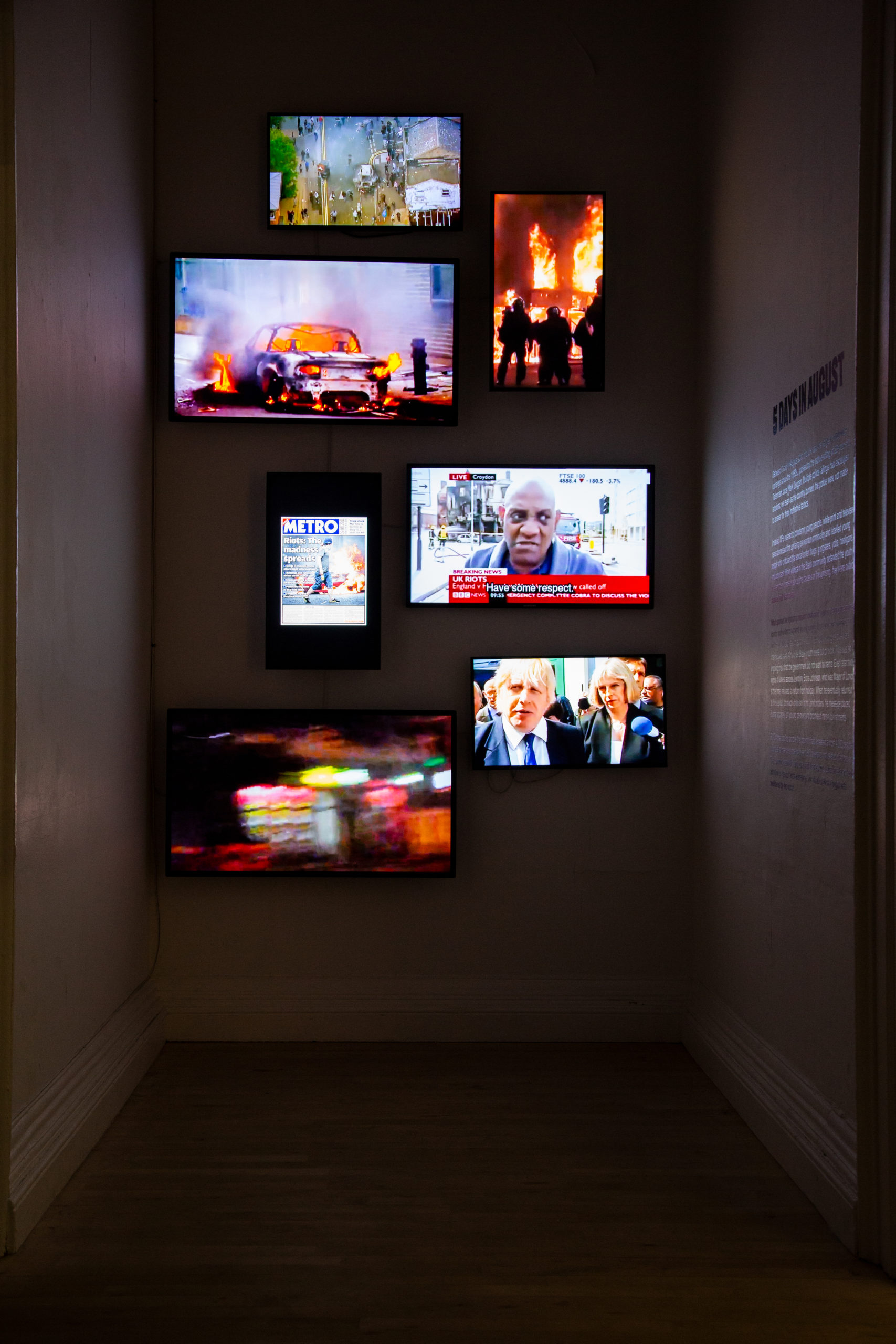 A dark room with 7 screens, displaying images of protests and news videos, mounted on the wall.