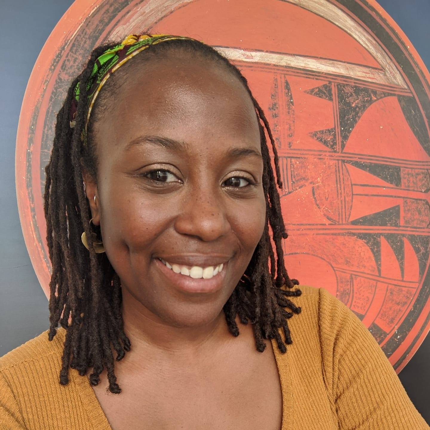 Jen is a Black woman with a warm smile, shoulder length locks, and a patterned head scarf. She is wearing a gold top, and stands in front of a black and red circular painting.