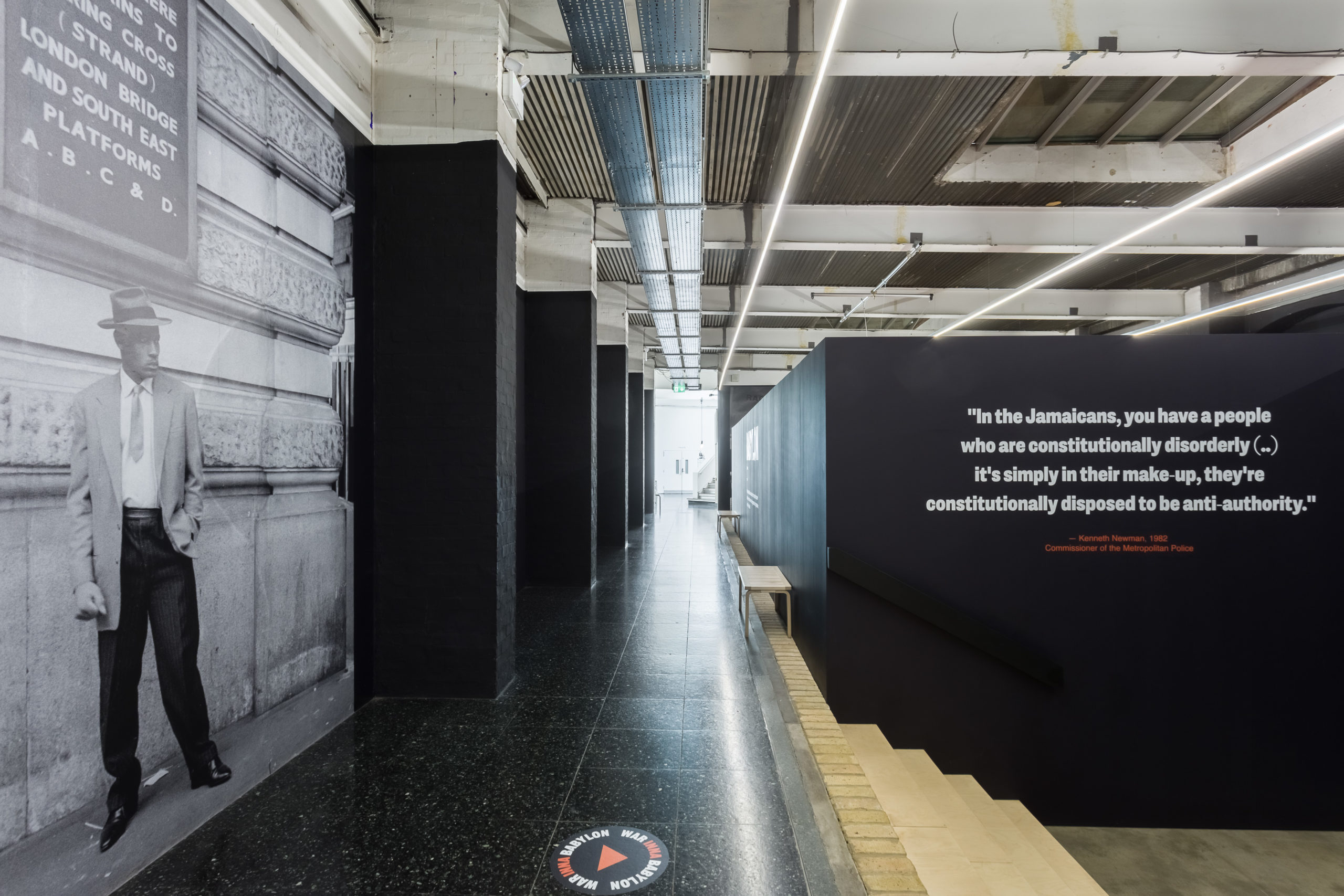 A hallway leading to an exit in the center, with a large photo on the left wall and a quote on the right wall next to steps.