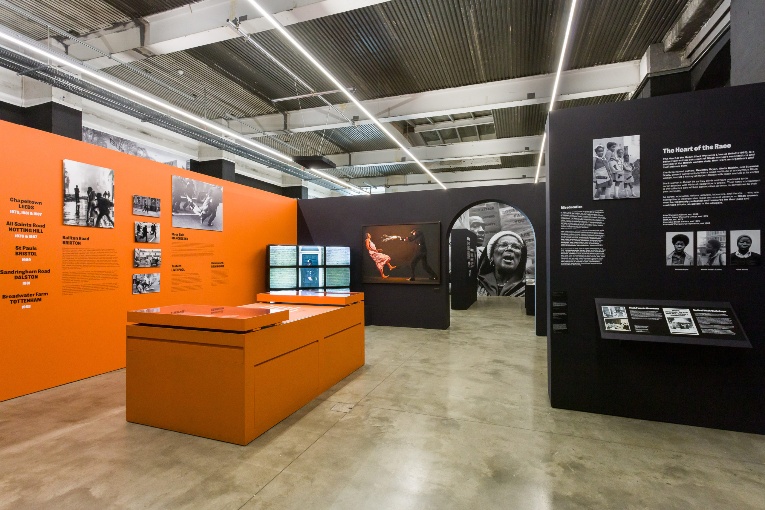A view of the exhibition featuring an orange wall and table with text and photos on the left and a set of black walls with photos and text on the right. An archway leads through one of the black walls in the center into another part of the exhibition.