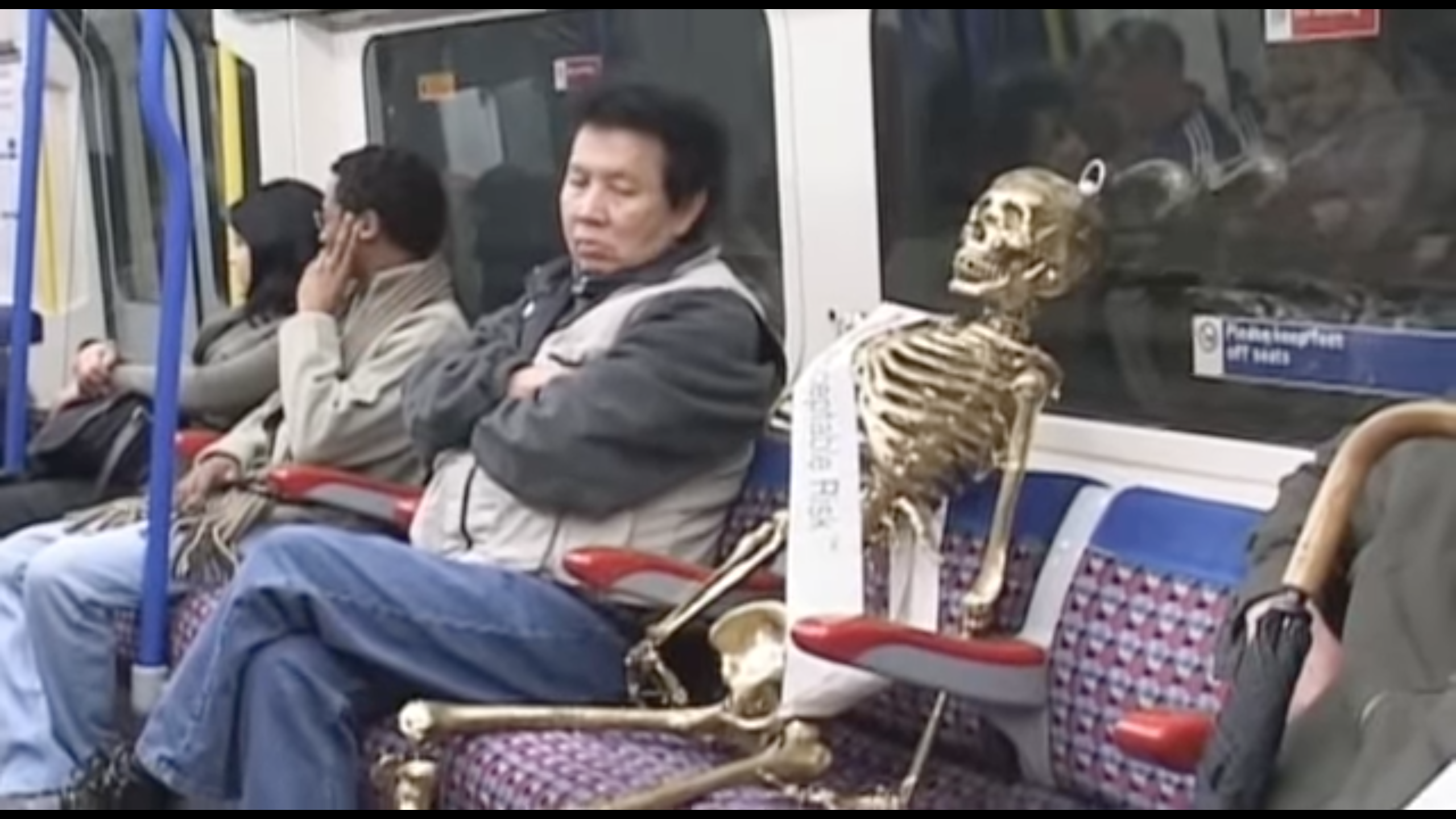 A screenshot in which a fake golden skeleton is seated on London public transit next to several people.