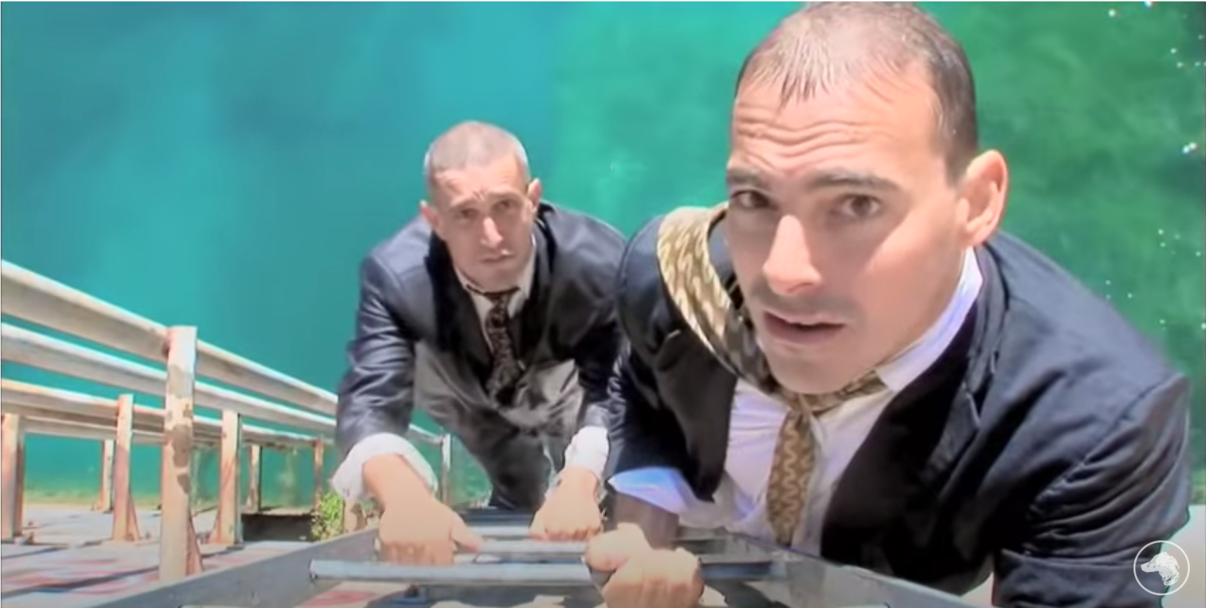 A screenshot of the Yes Men looking up at the camera and wearing suits as they climb up out of water on a ladder.