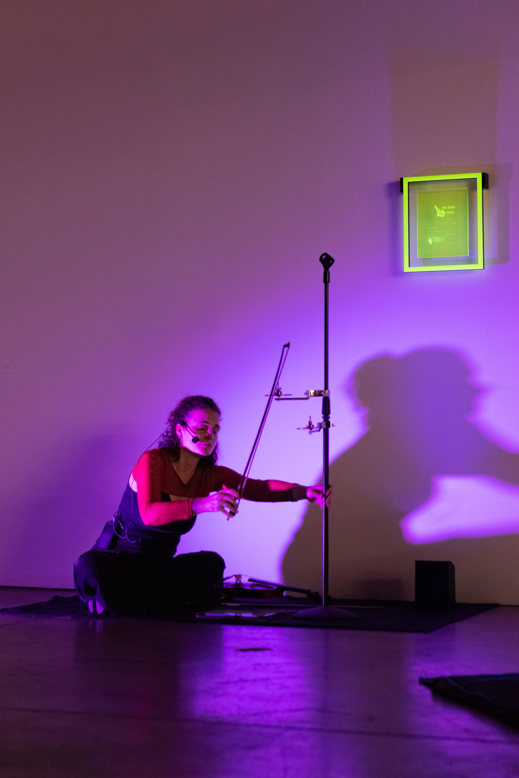 A woman seated on the ground takes a violin bow against a small symbal, her shadow cast behind her and a rectangular piece of translucent yellow acrylic hanging above it.