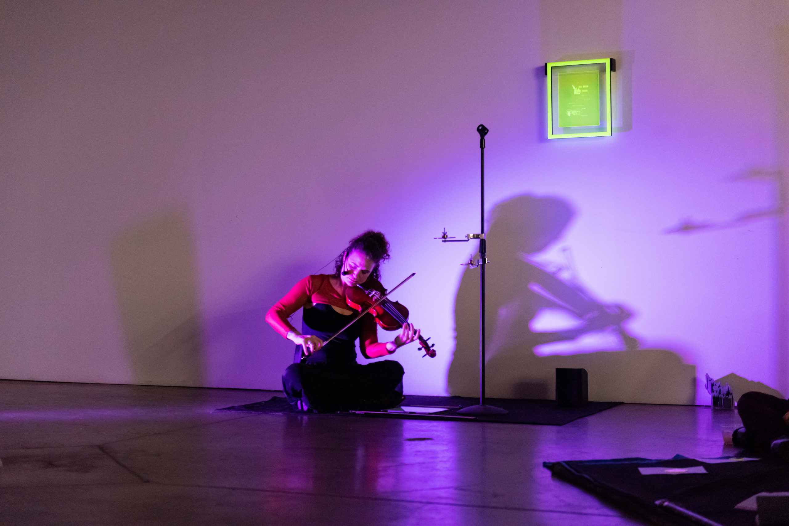 A woman seated on the ground plays the violin, her shadow cast behind her and a rectangular piece of translucent yellow acrylic hanging above it.