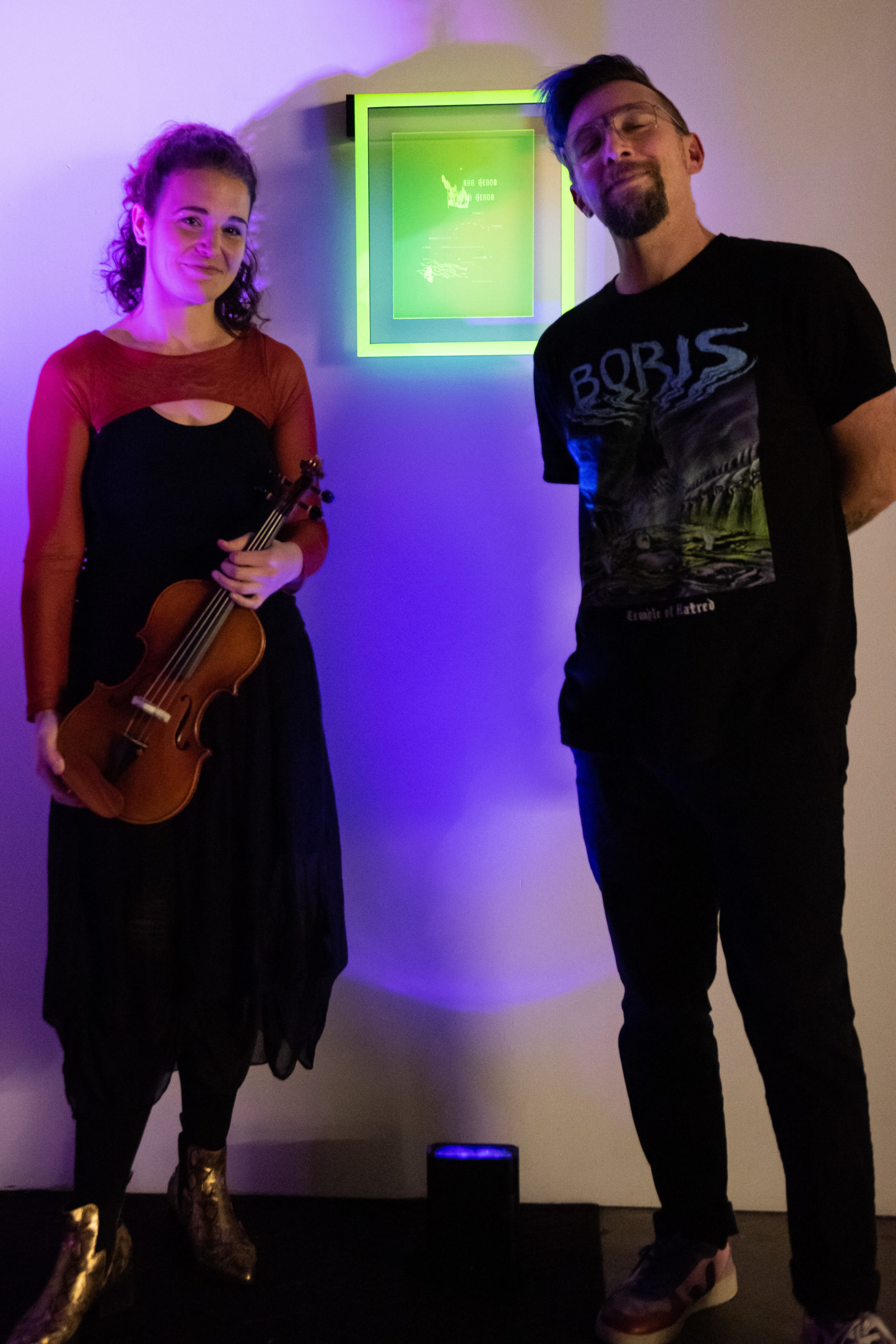 Two artists, one holding a violin, the other hands behind his back stand on either side of their collaborative piece of neon glowing yellow acrylic