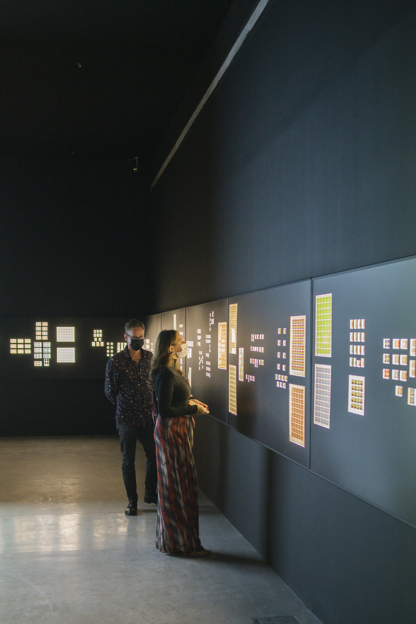 Two people view square and rectangular grids of stamps, backlit amongst a series of panels that span across the walls.