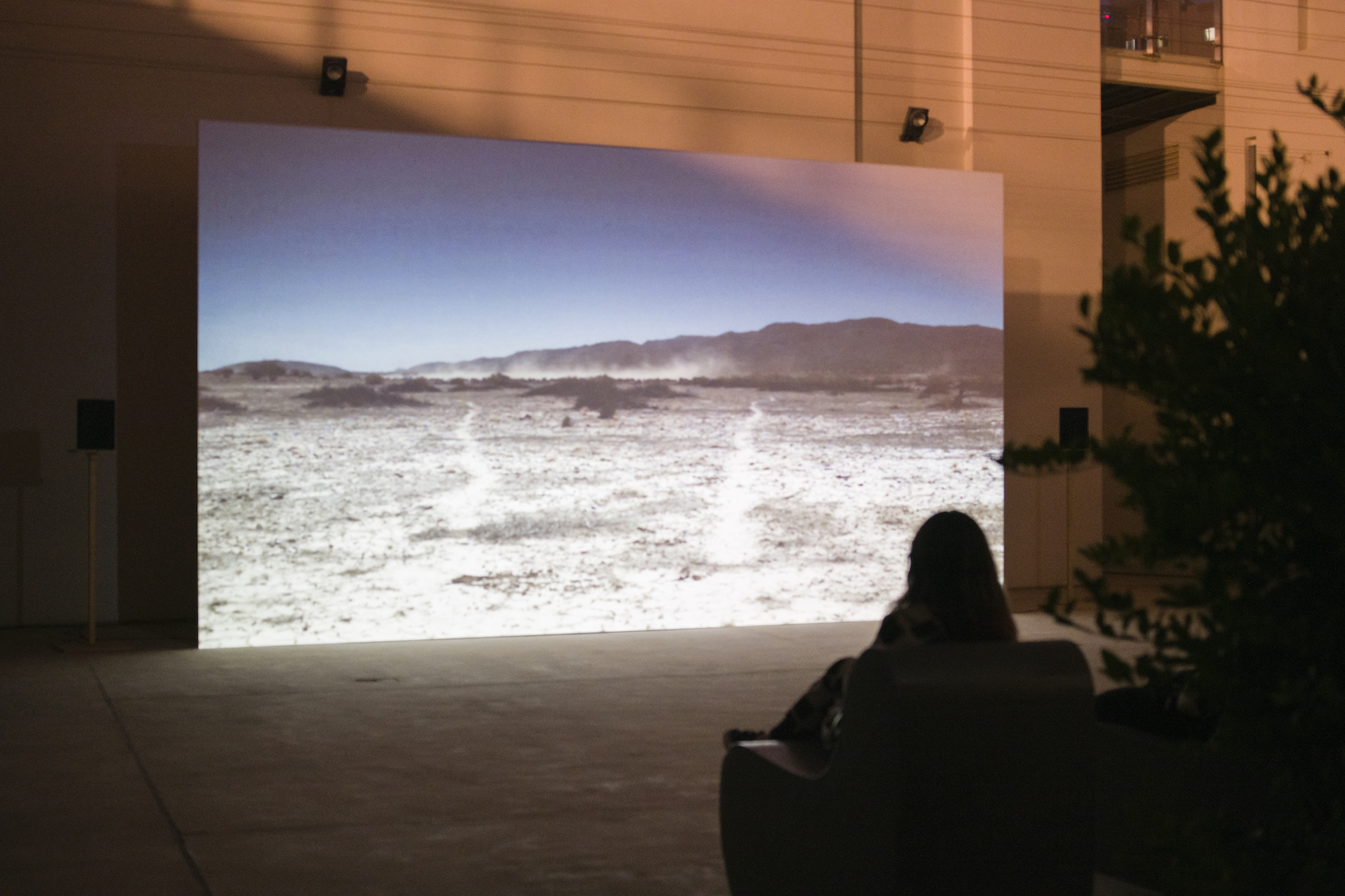The silhouette of a figure sits in a chair in the foreground before a screen depicting a desert landscape sits in the background amongst an urban space.