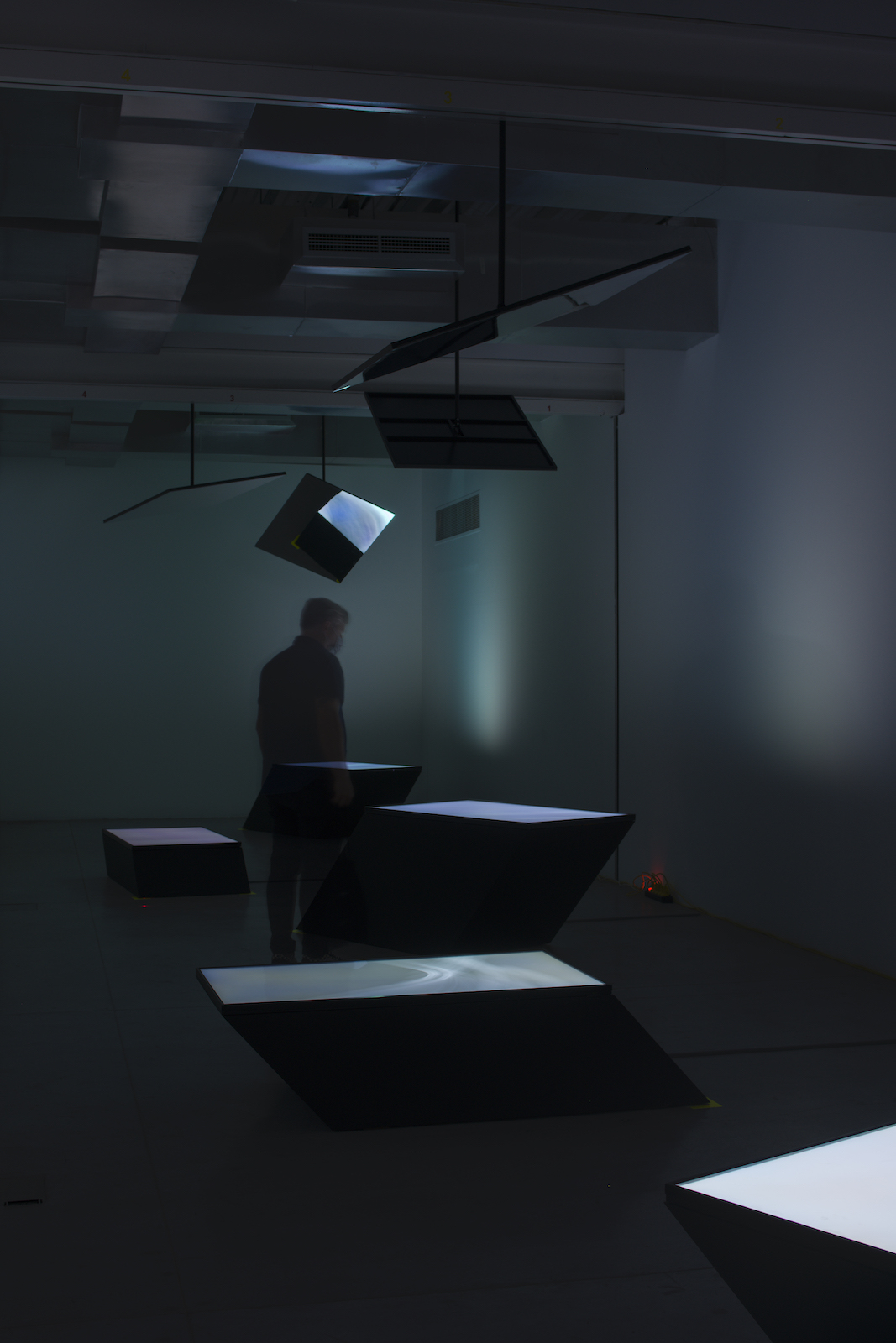 In a dark room, a figure stands among a number of screens facing upwards, shining a pale light as they reflect off four screens or mirrors angled down from the ceiling.