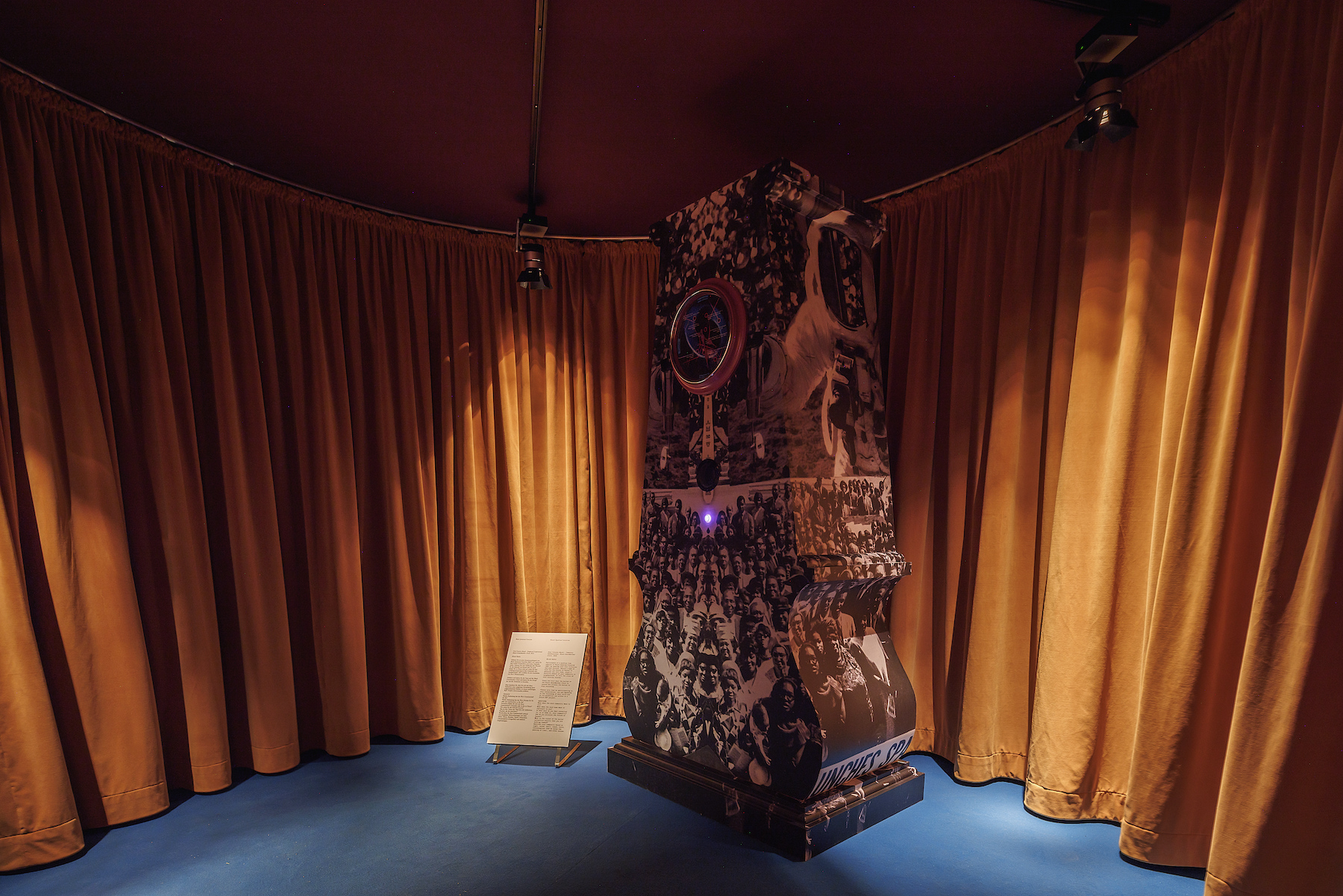 A big grandfather clock with photographs of different people printed on the clock is in the middle of a dark room with dark yellow curtains and blue flooring.