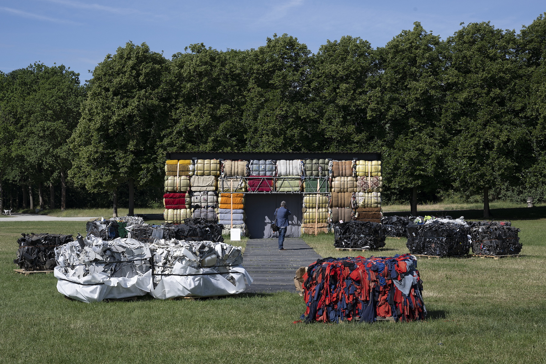 Big colorful blocks of fabric, paper, and plastic are grouped together by color in an outdoor space, with a grey walkway leading to big blocks of fabric installed onto a metal frame.