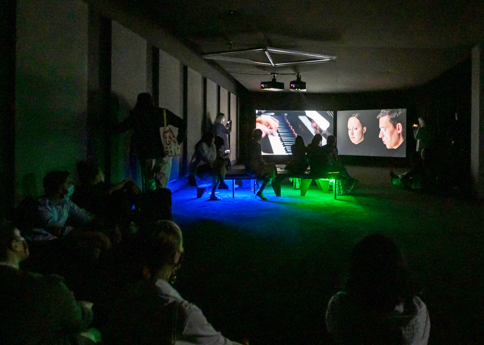 A group of people sitting around a dark room, focused on two large screens featuring piano players and hands on the keys are projected in the background.