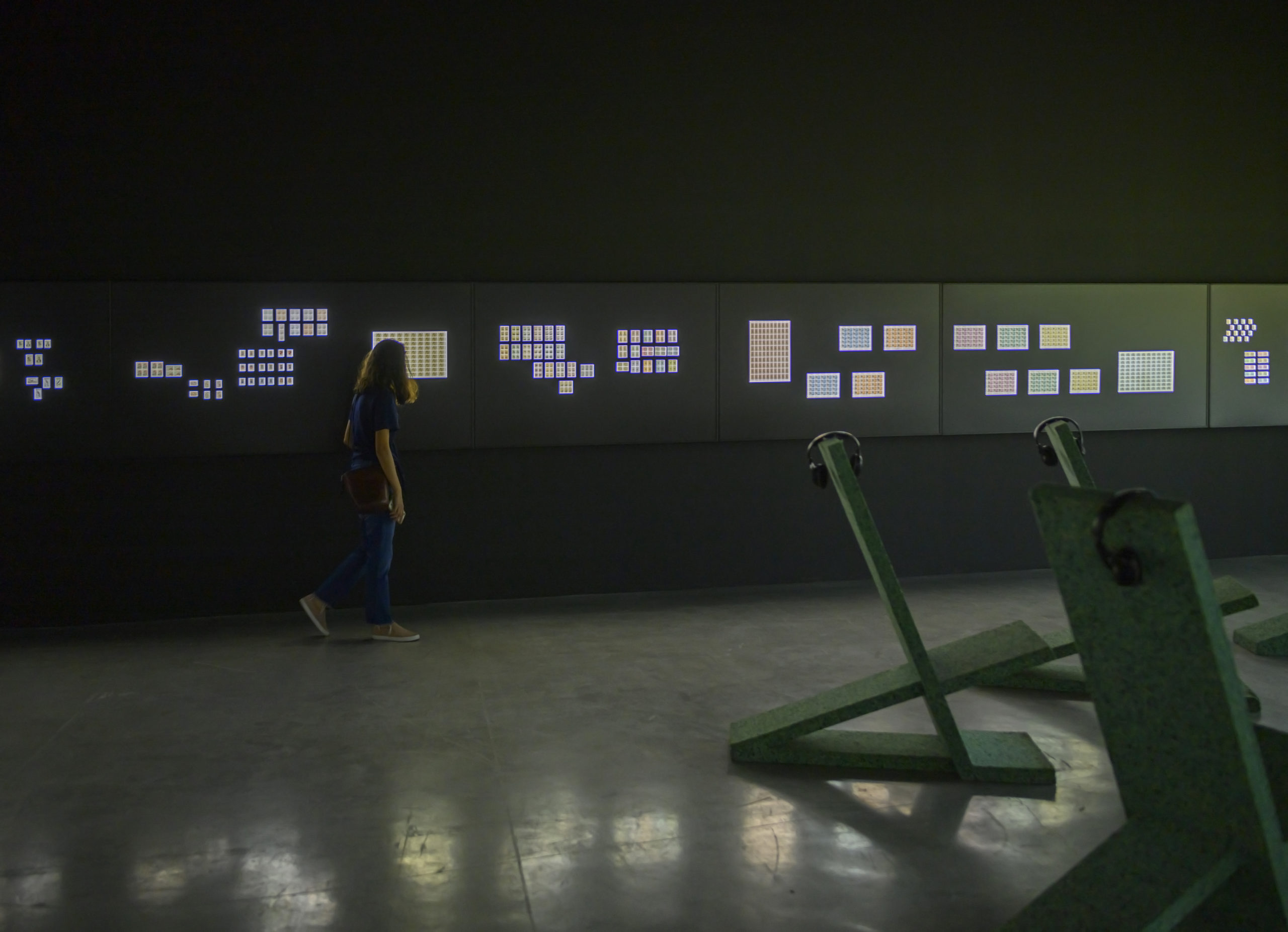 A person walks along a series of panels containing a number of illuminated stamps with a series of chairs and headphones reside in the foreground.