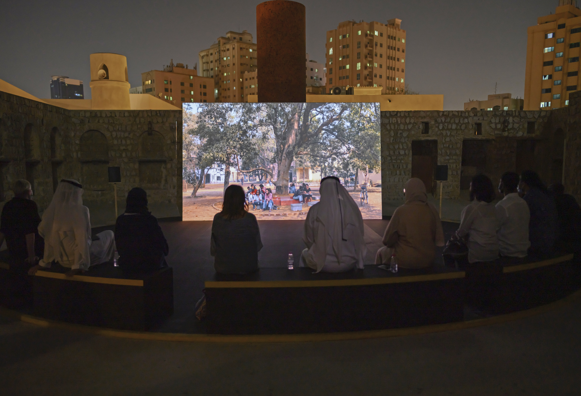 A group of people sit in a semi-circle in the foreground in a courtyard, viewing a move on a screen in the center depicting a group of people around a tree.