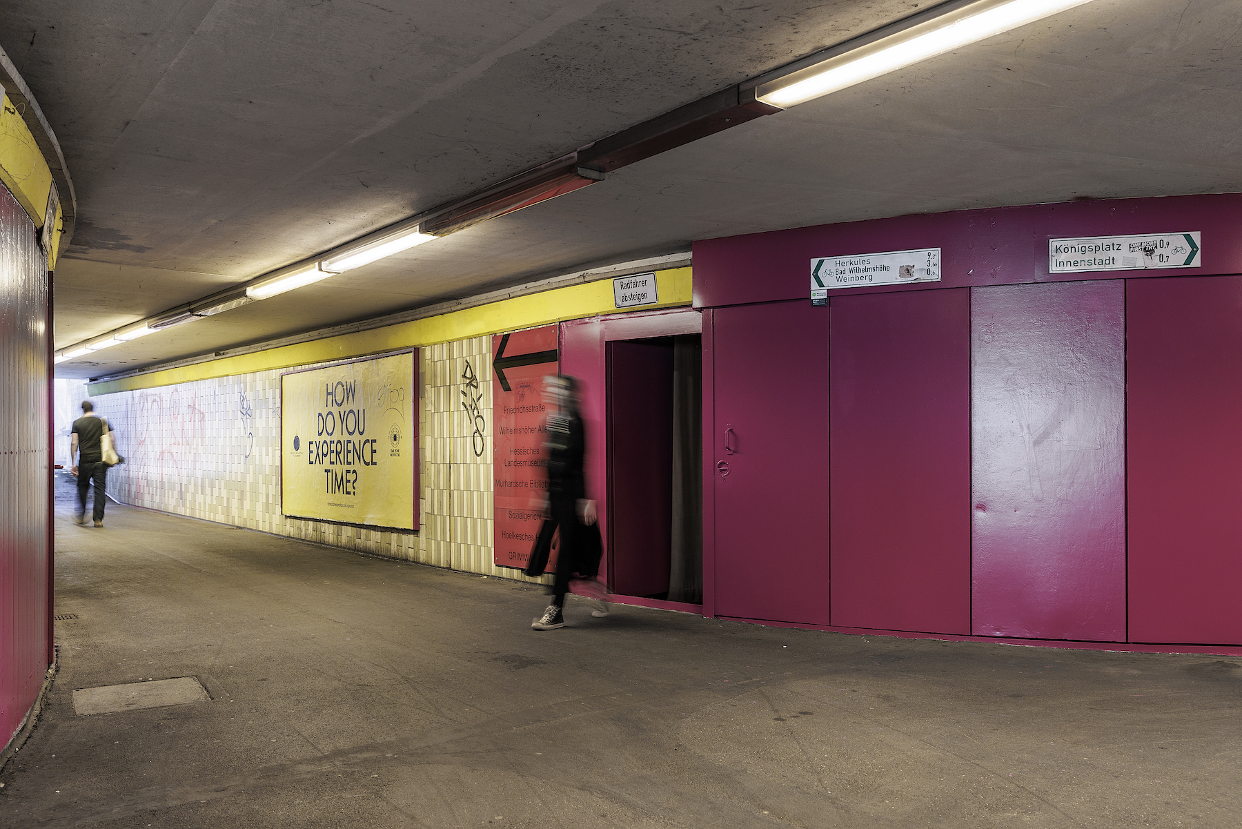 A long hallway in a subway station with pink and cream colored walls, with a sign that reads, 