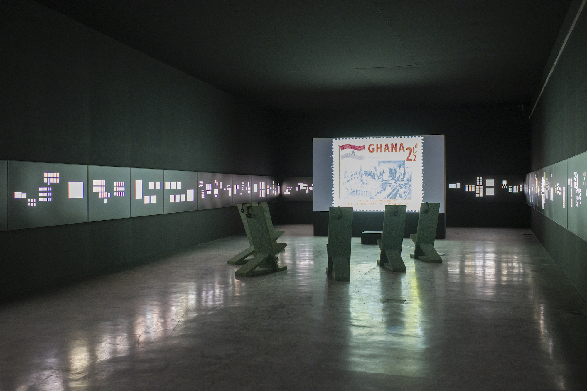 A room exists with panels of stamps on its walls and a large screen depicting a stamp reading Ghana is at the center of the room surrounded by chairs.