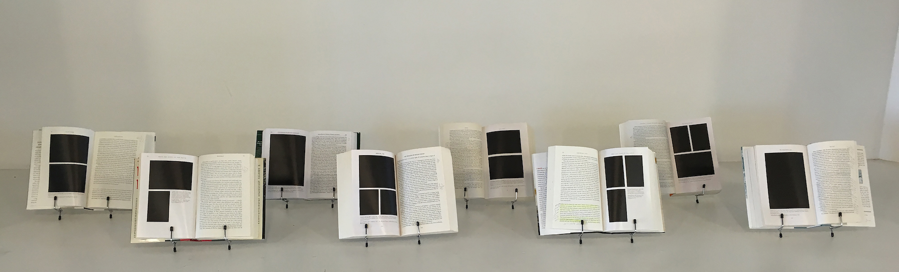 An installation view of eight copies of the same book opened to different pages, displaying different blacked out photographs and text.