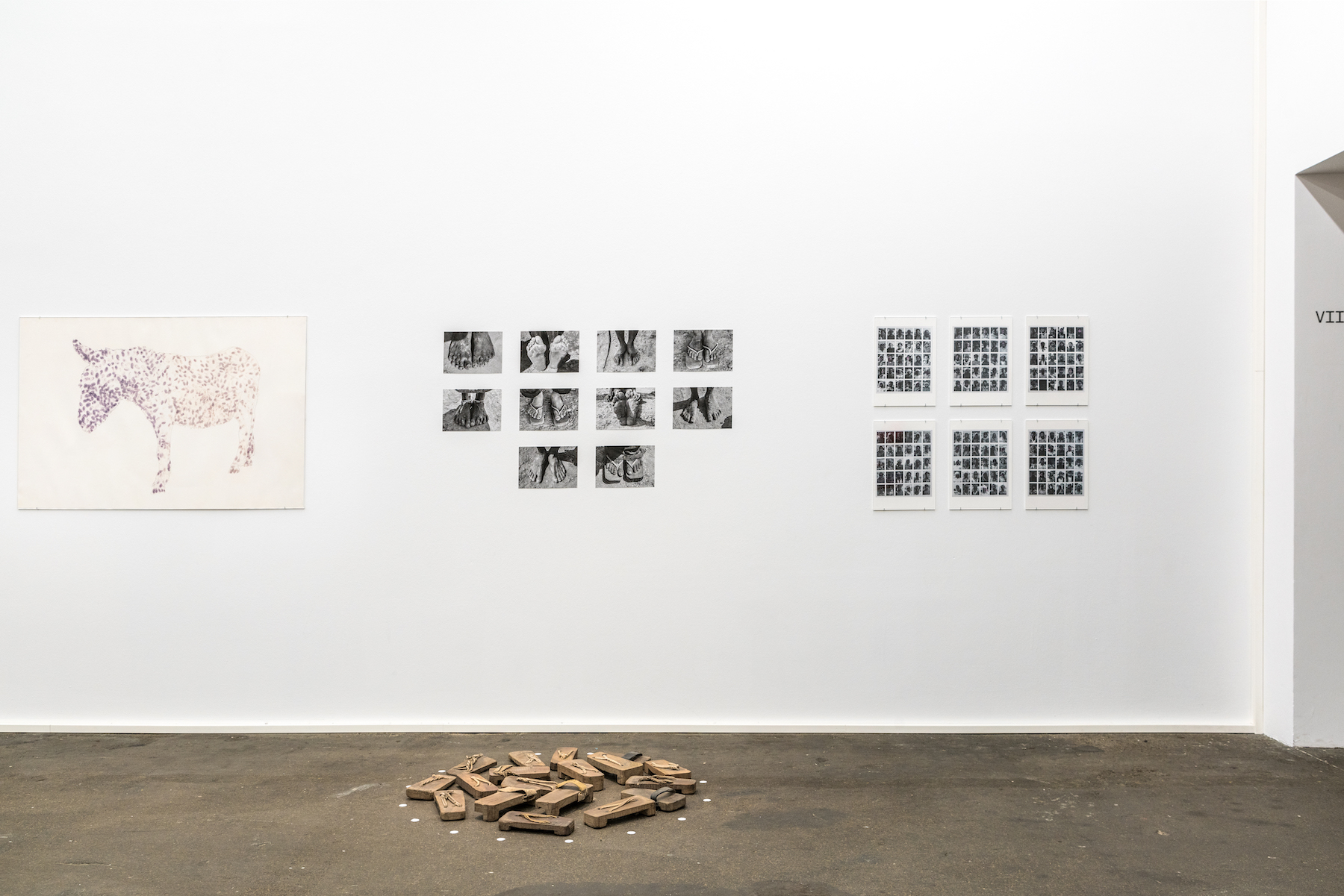 From left to right, a donkey, ten groupings of photos of feet, and six different groups of smaller black and white images are displayed on the wall. On the floor in the middle, a circle of rectangular wooden sandals are displayed.