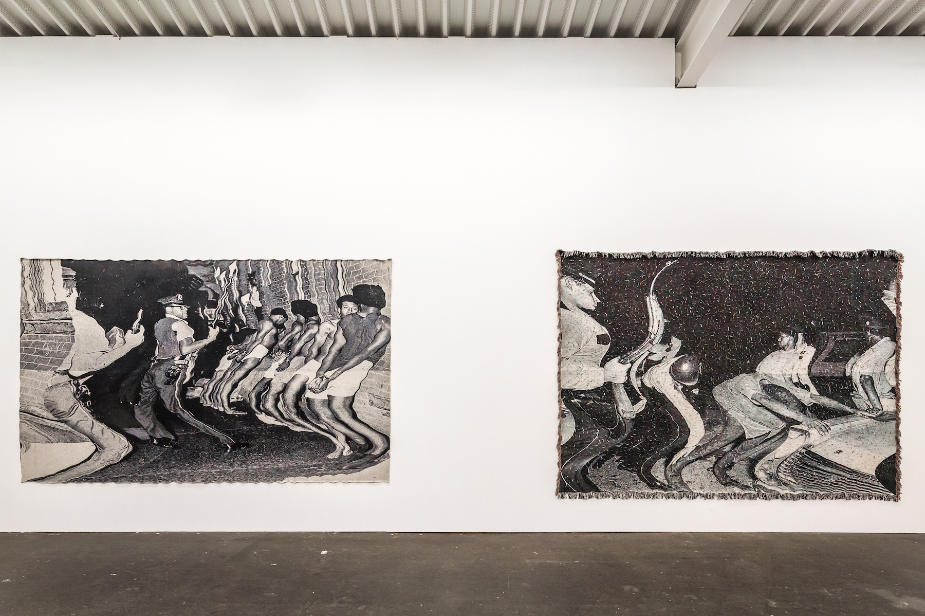Two black and white tapestries with printed black and white photos of police violence are displayed on a white wall.