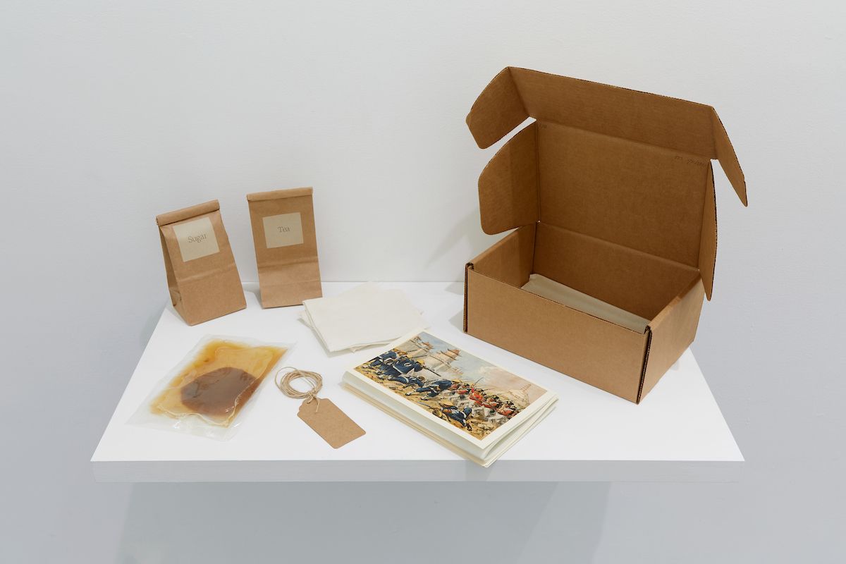 A white shelf containing various items from left to right; two teabags, a compressed container with teabag in center, teabag labels, a postcard of colonist images, white napkins, and an open cardboard box.