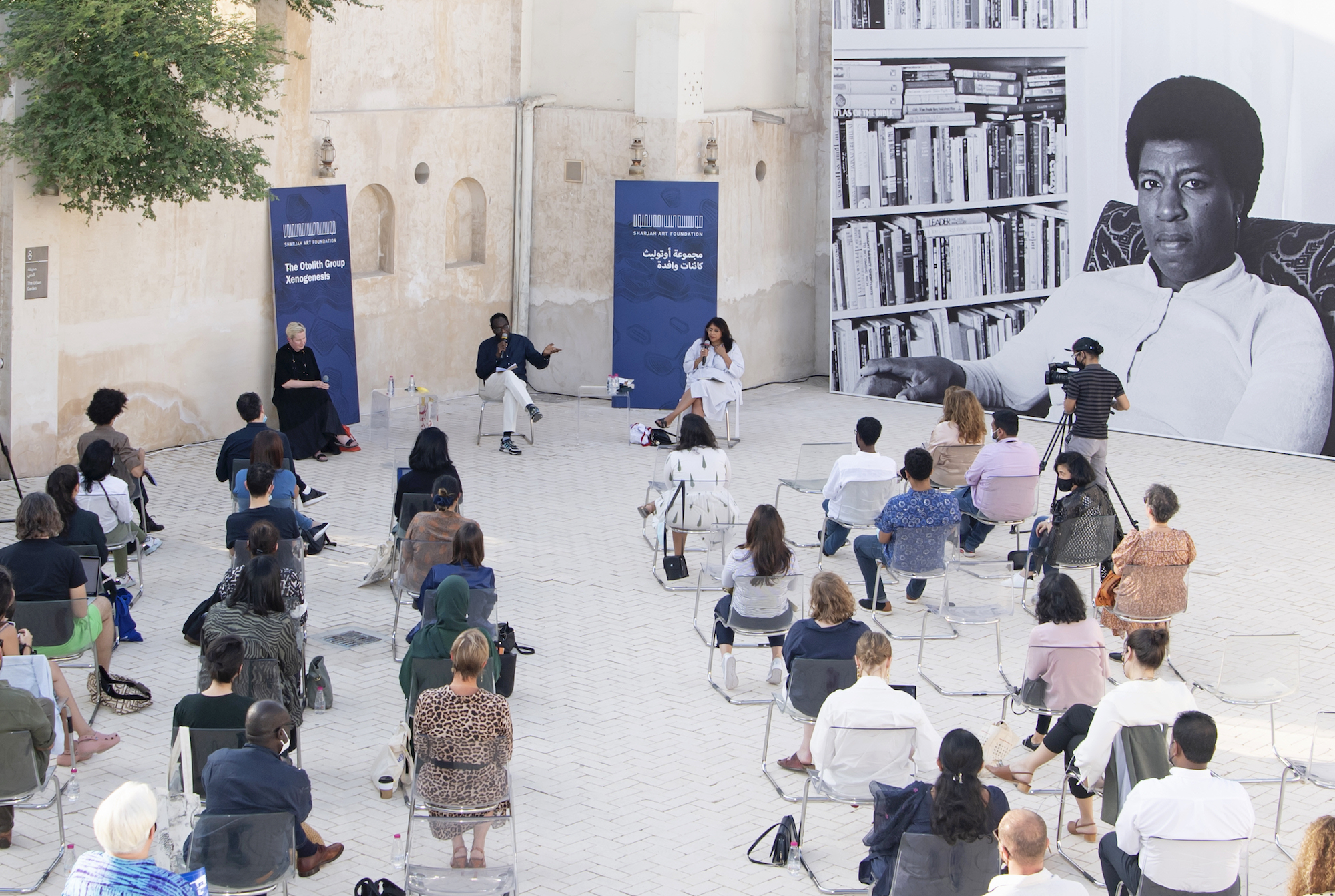 In a brightly lit coutryard during the day, three people sit in chairs with microphones as a crowd of people sitting in their own chairs listens before them, attentive and facing them.