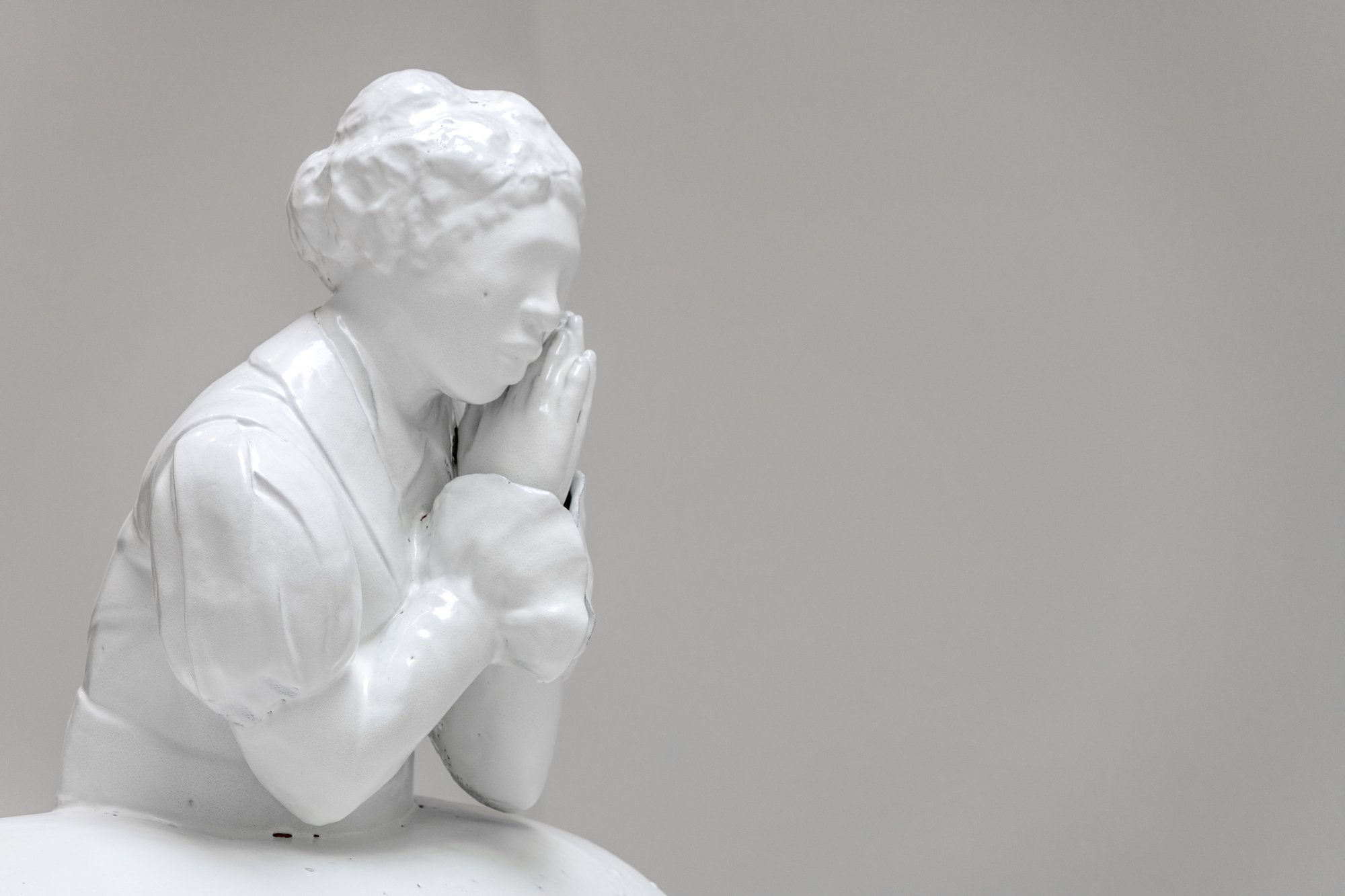A white ceramic piece of a woman with hands clasped together resting beside her face faces outward with all details left blank; a stark white statue.