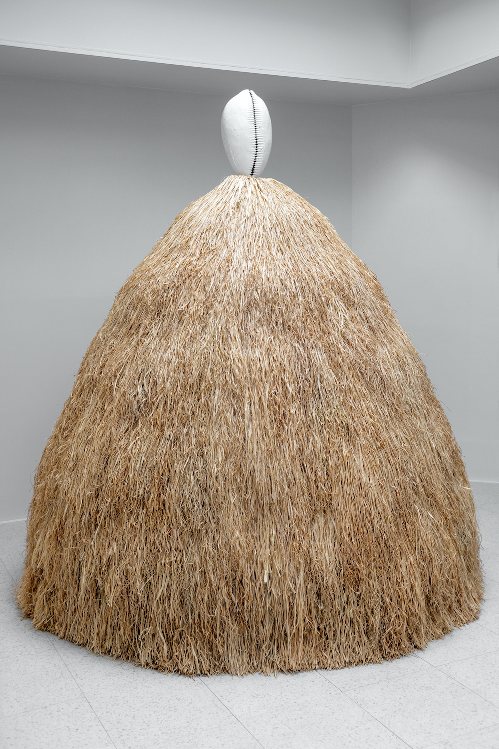 A large sculpture of straw colored raffia resembling a skirt leads up to a stoneware piece of ceramic resembling a shell amidst a stark white room.