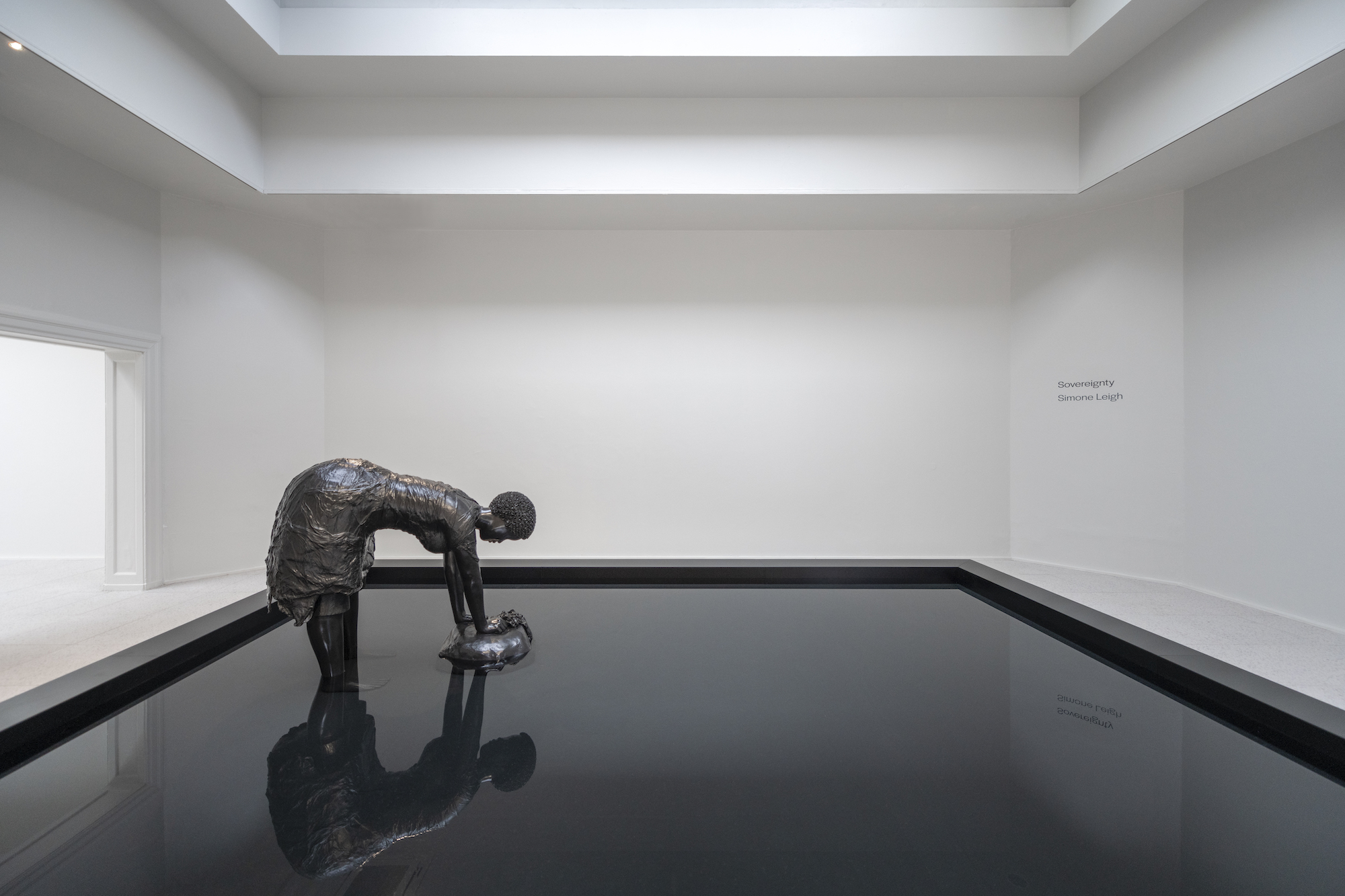 A dark bronze sculpture of a woman bent over a pool of water, her image being reflected.