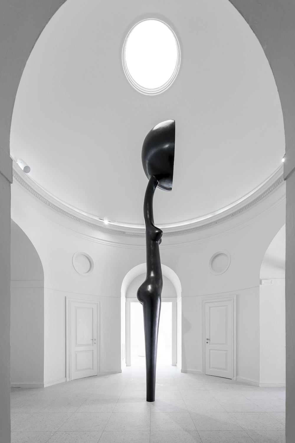 The side of a tall black bronze statue of an abstracted figure with a large head stands erect in a stark white room.