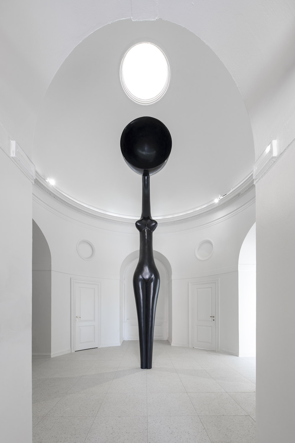 The front of a tall black bronze statue of an abstracted figure with a large head stands erect in a stark white room.