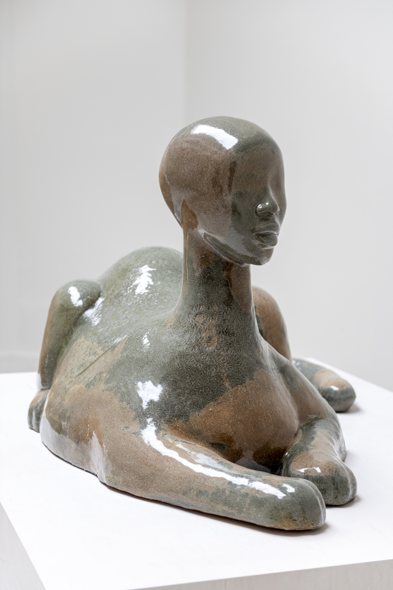 A shiny brown / green sculpture of a sphinx laying down atop a white pedestal.
