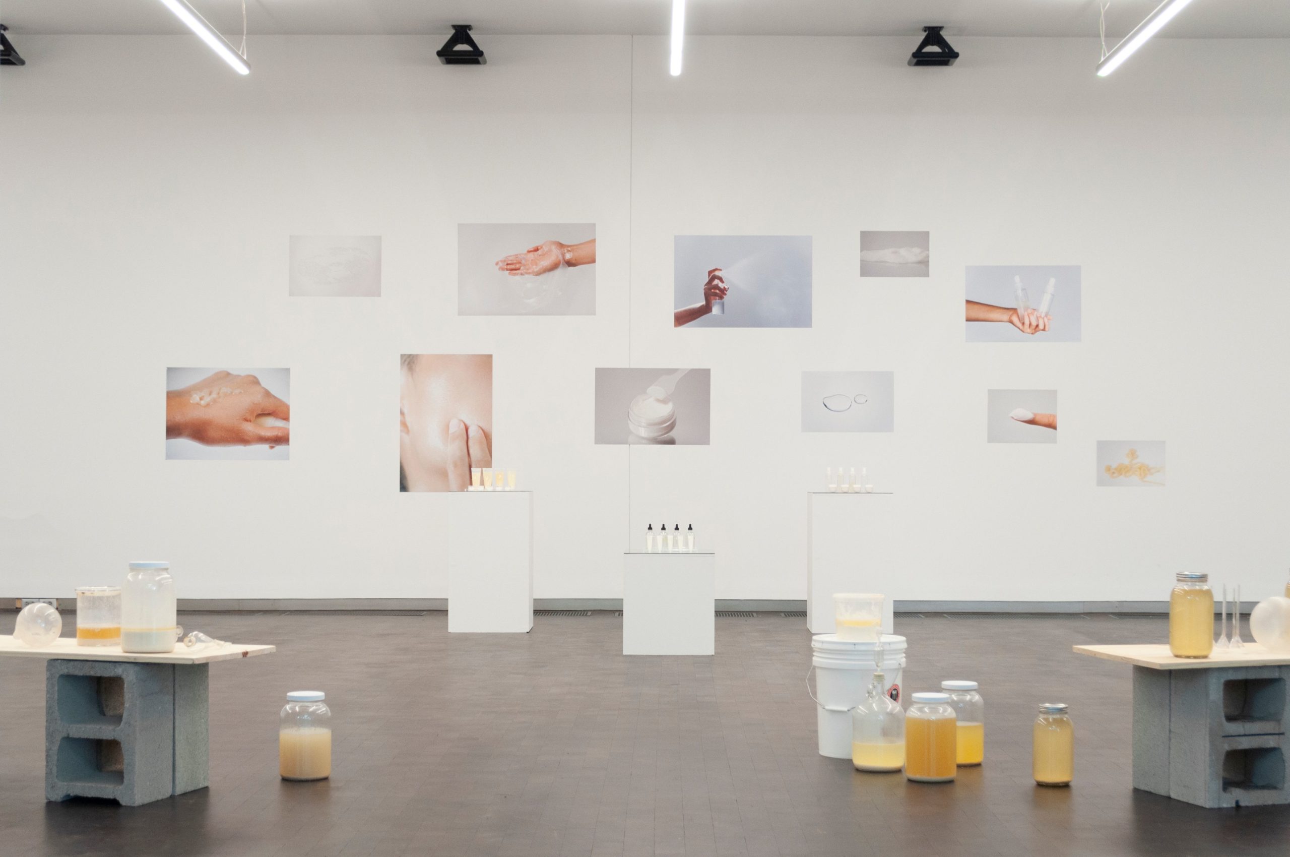 A shot of a room with various canisters filled with yellowish liquids displayed on cement blocks in various shelf sizes, with images of hands and skin holding bottles and spraying foam on the far wall.
