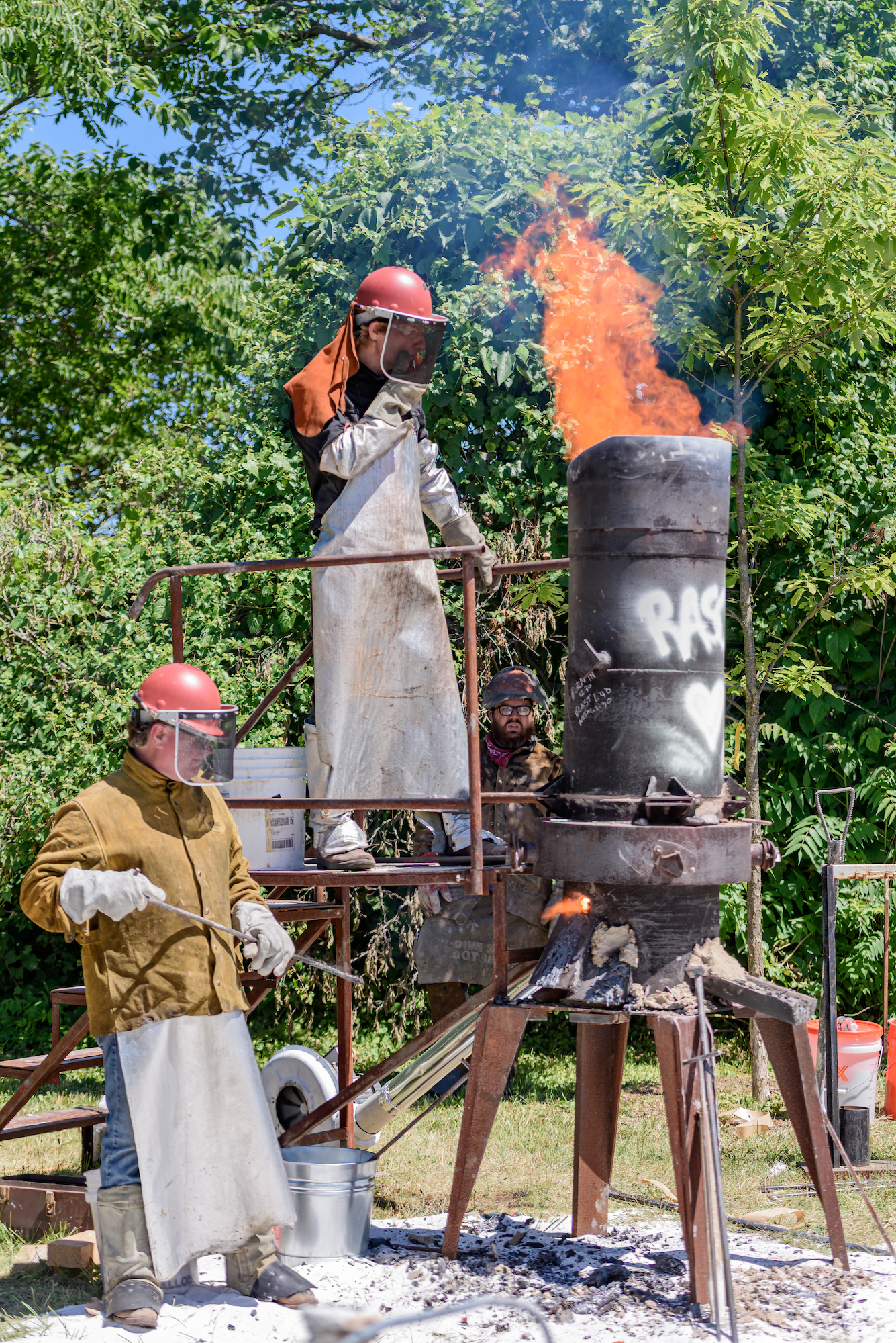 Two people stand before a large cylindrical furnace spewing fire from the top