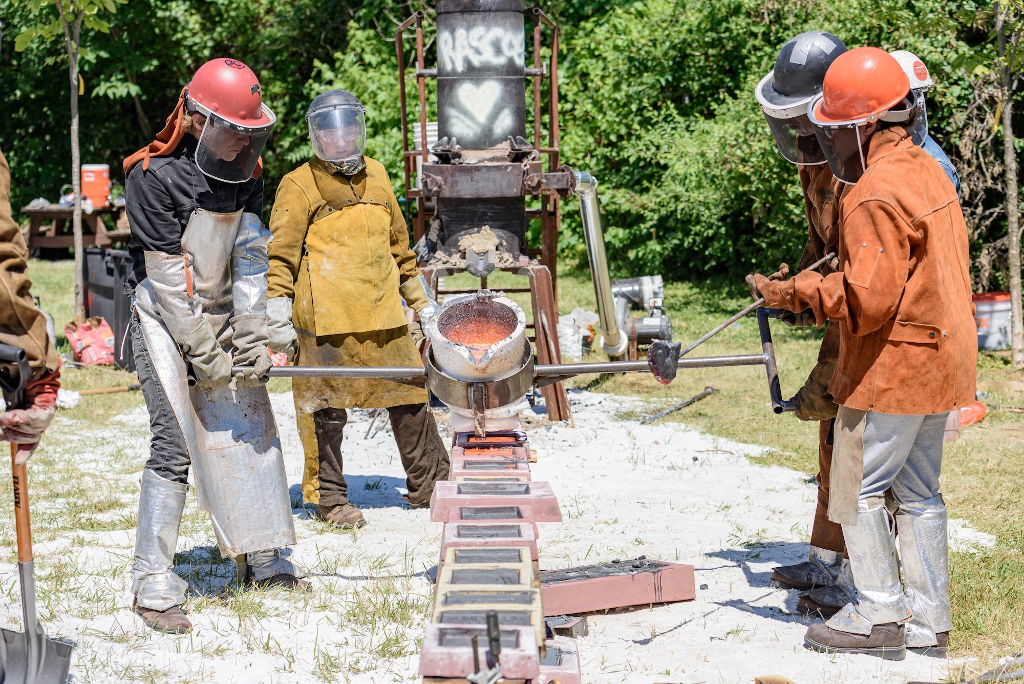 Two figured balance a crucible from which molten iron is being poured in the bright hot summer sun against a verdant green background