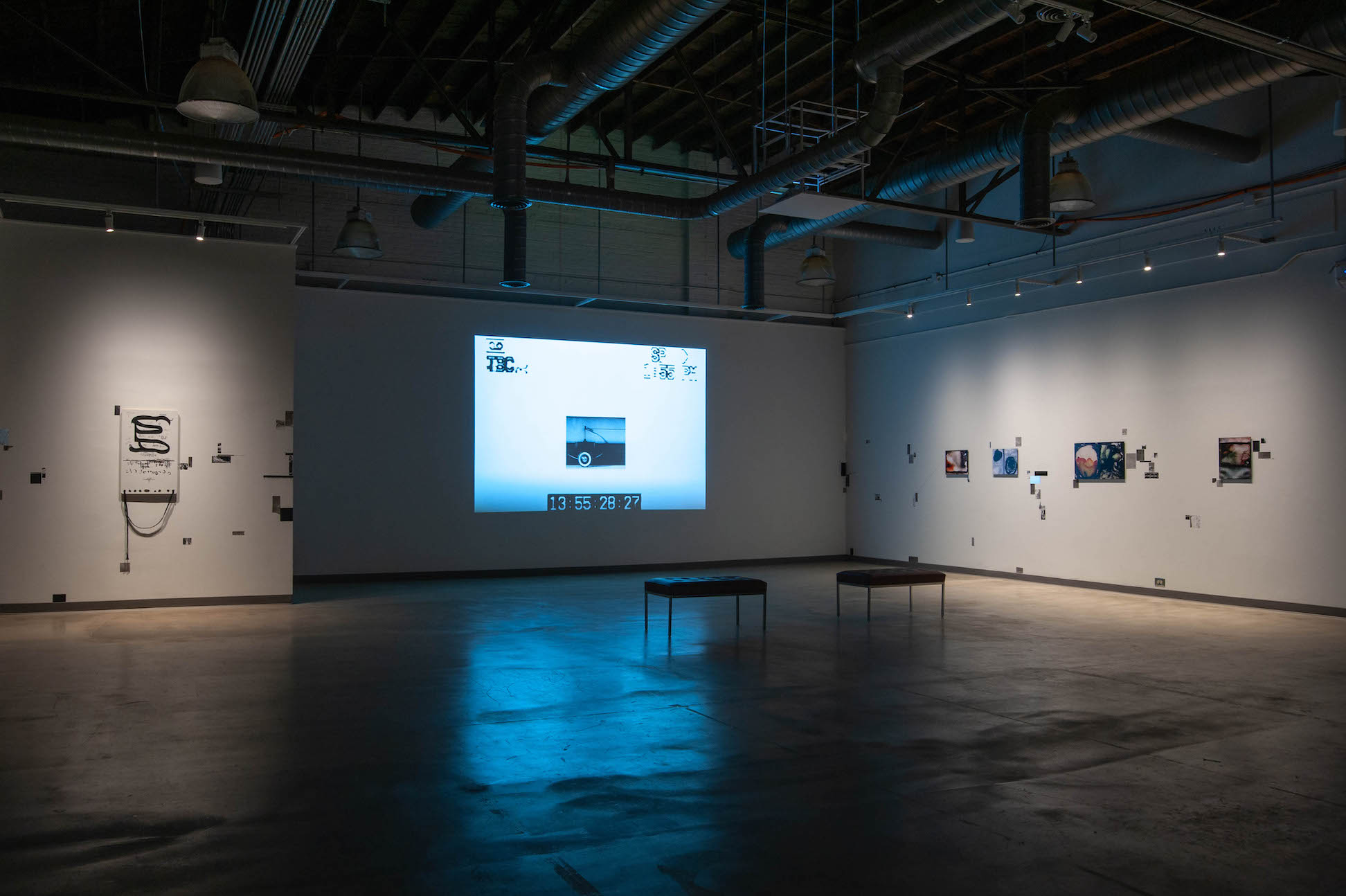 A view of the exhibition space, with two leather benches in front of a big screen projector. On both sides of the projector on the other walls, there are different artworks and text.