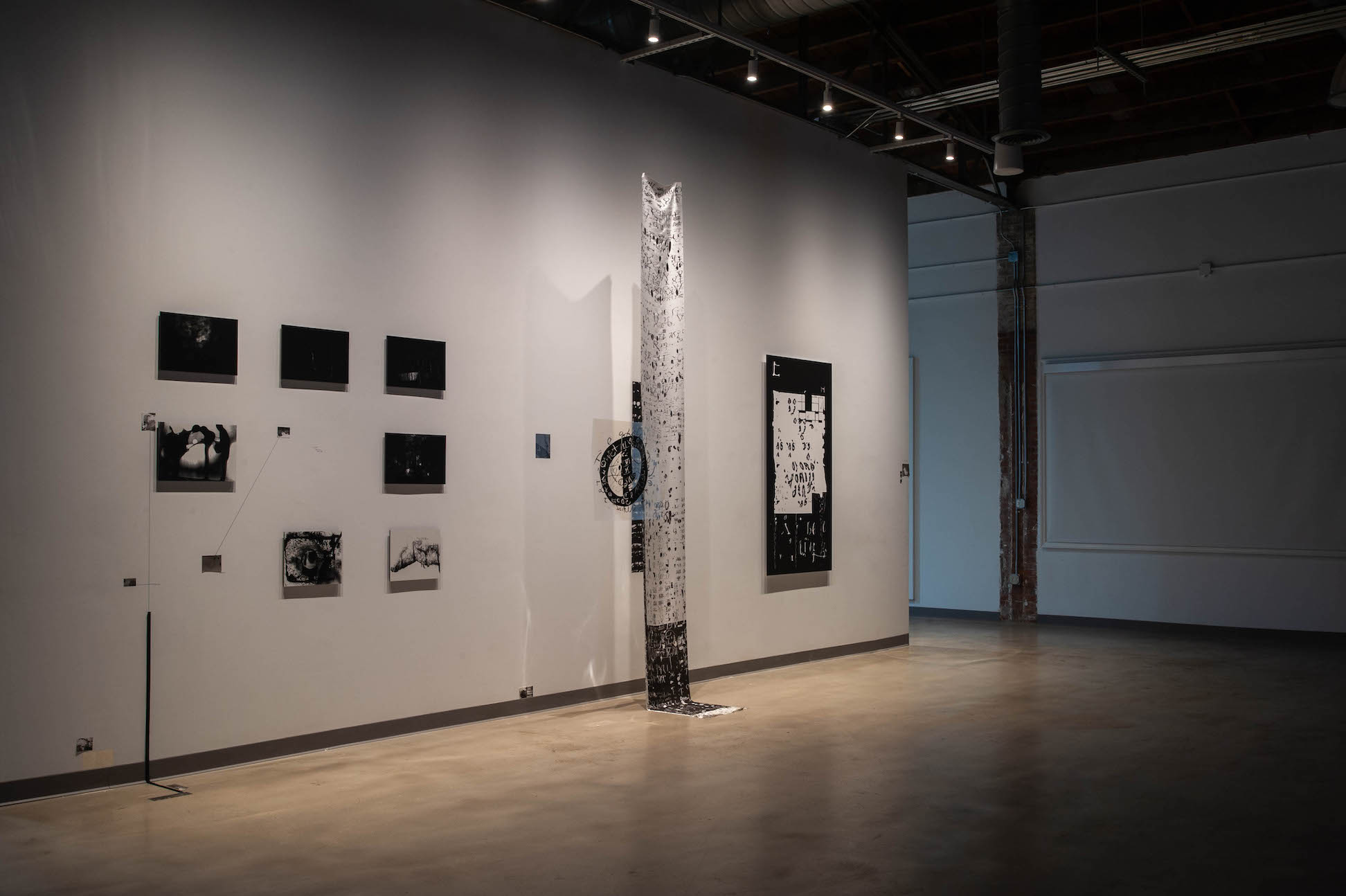 An exhibition view of one wall, with a tall narrow black and white column in the center, with groupings of black and white images on either side of the column.