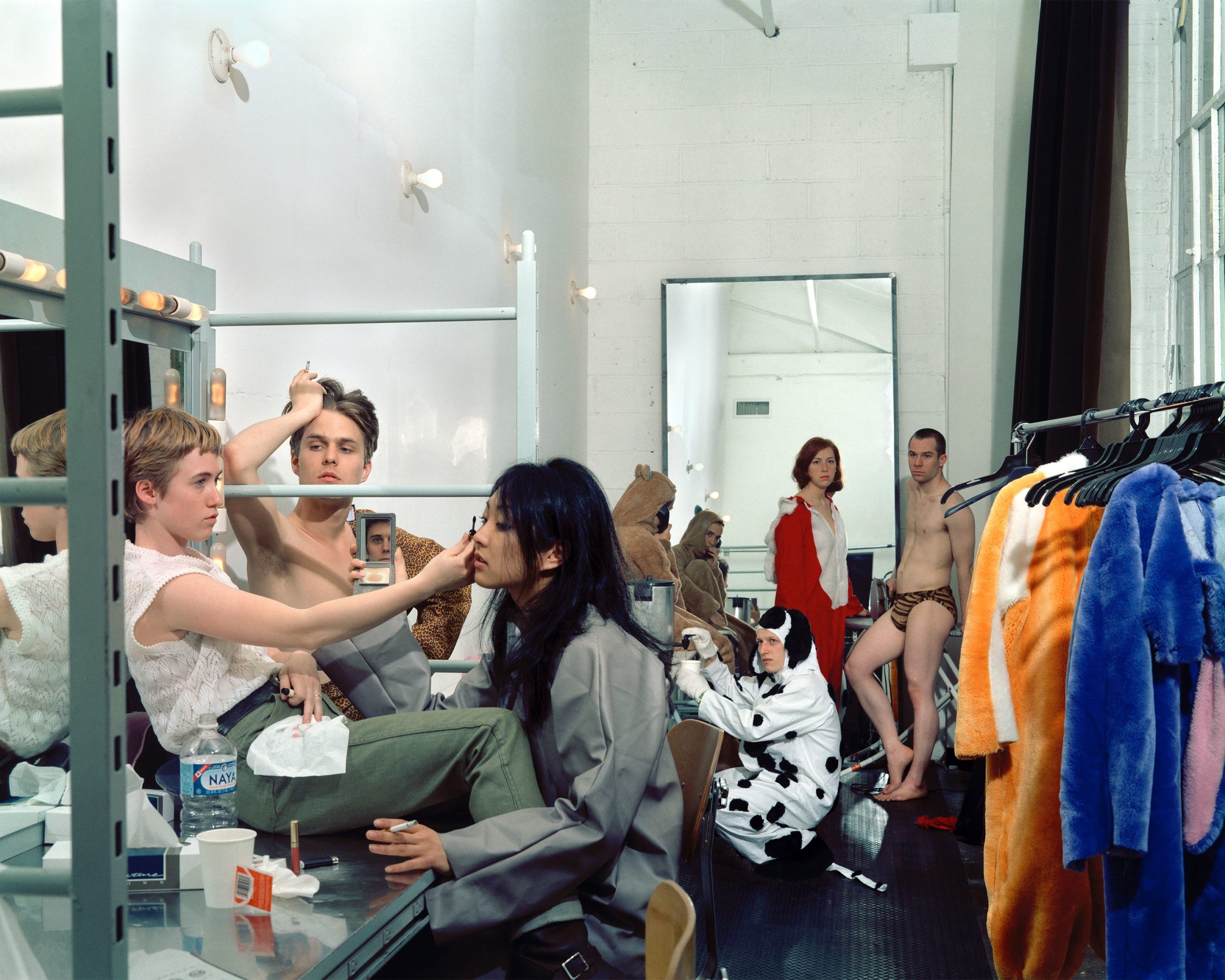 7 people stand in a makeup or dressing studio. Two women at the forefront appear to be applying makeup, with one of the women staring at her reflection by the mirror provided by her counterpart. A man watches behind them. Four figures in the background dress in animal suits, self-consciously looking towards the camera.