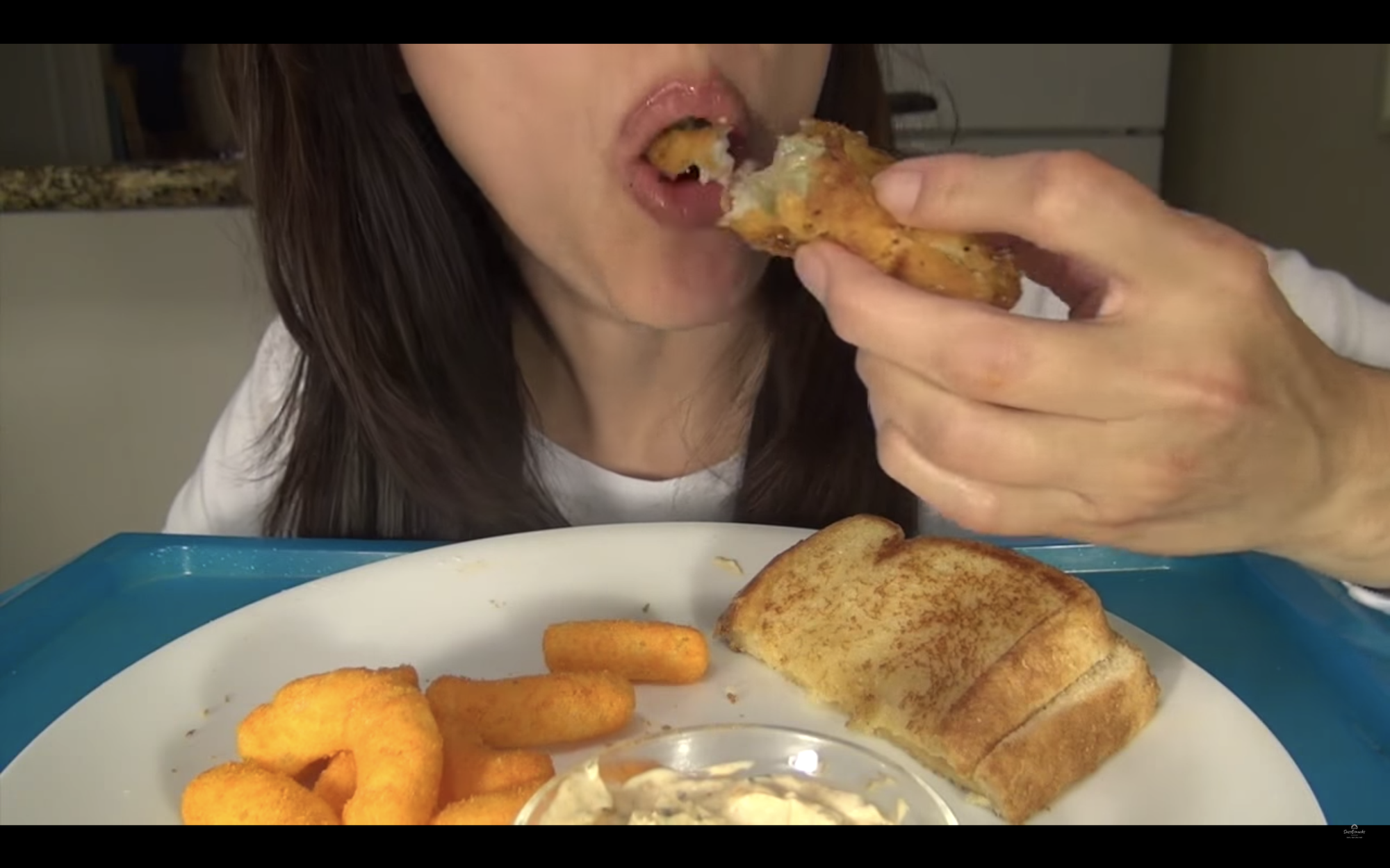 A close up video of a woman eating a fried pickle, half of it in her mouth, the other half held in her hand with a plate of cheese puff and half of a grilled cheese.