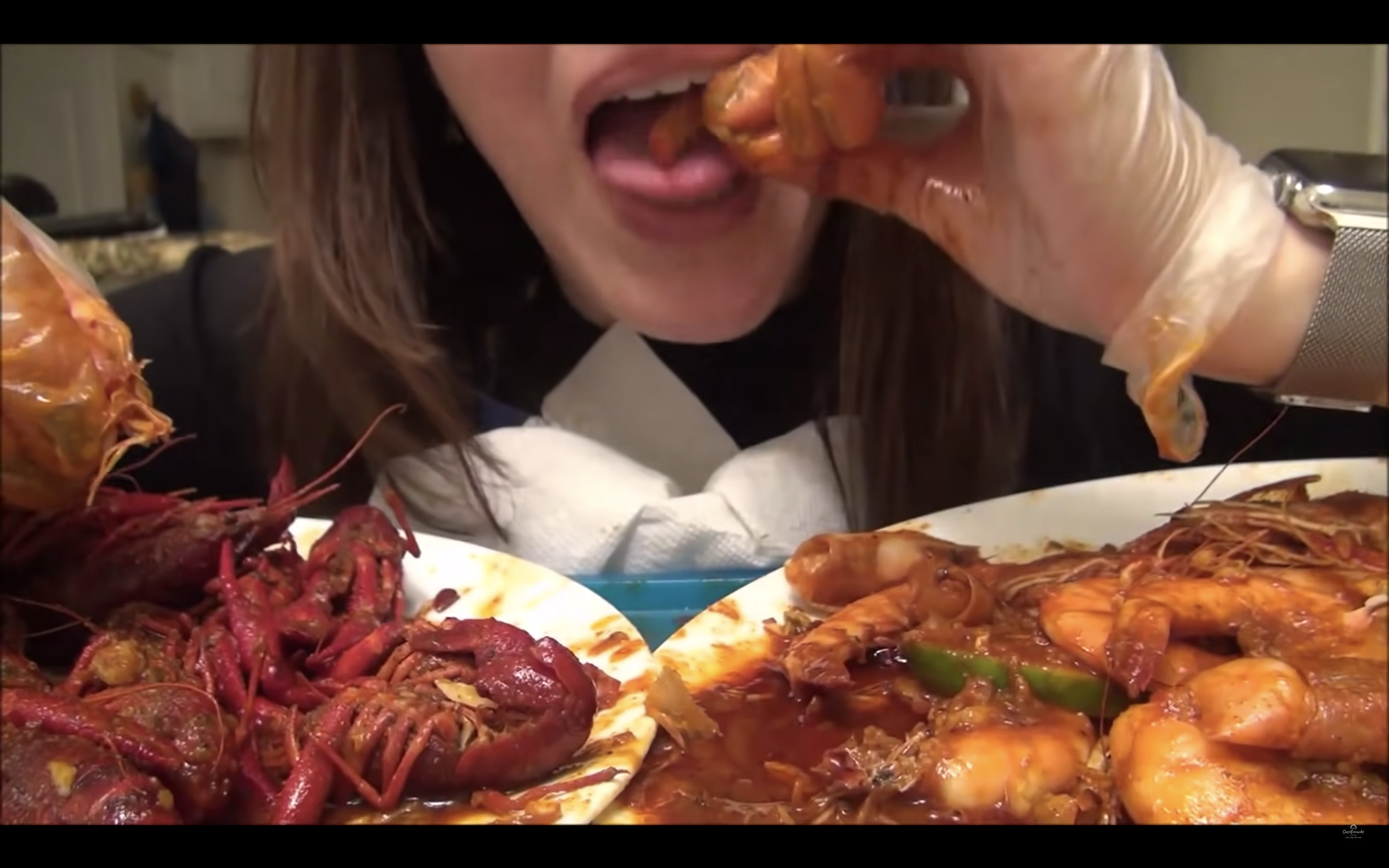 A person wearing latex gloves is placing a crawfish in their mouth and are about to bite down as there lay two plates of red and saucy